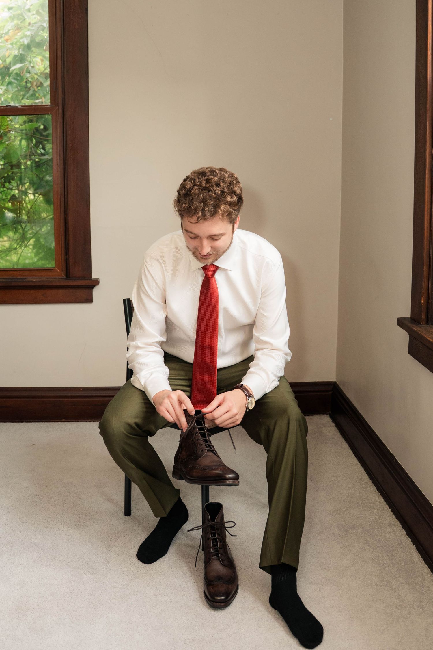 Groom in white shirt and red tie lacing brown leather boots while seated on chair before wedding.