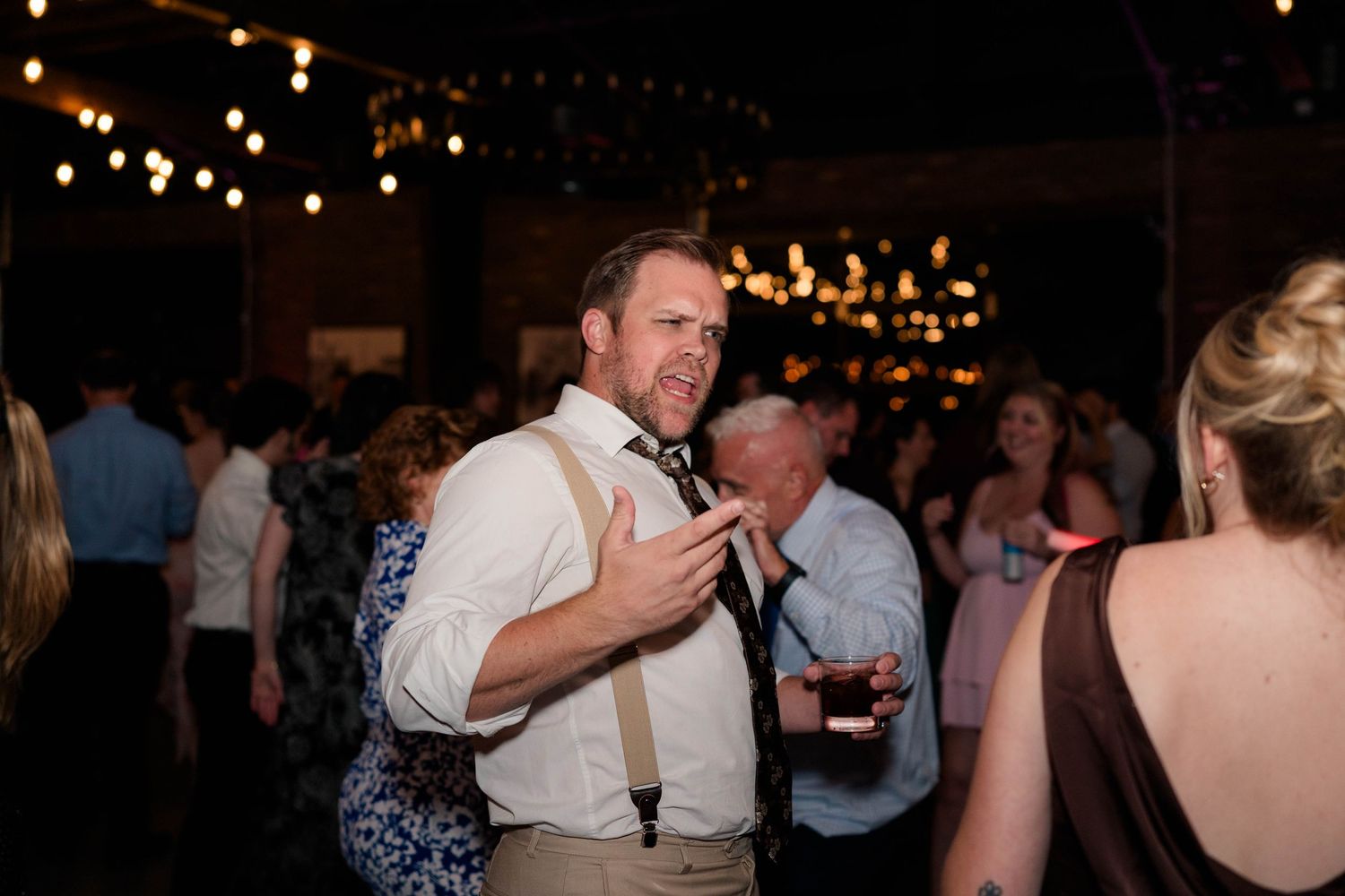 Energetic wedding guest dances with suspenders and tie at lively reception under string lights.