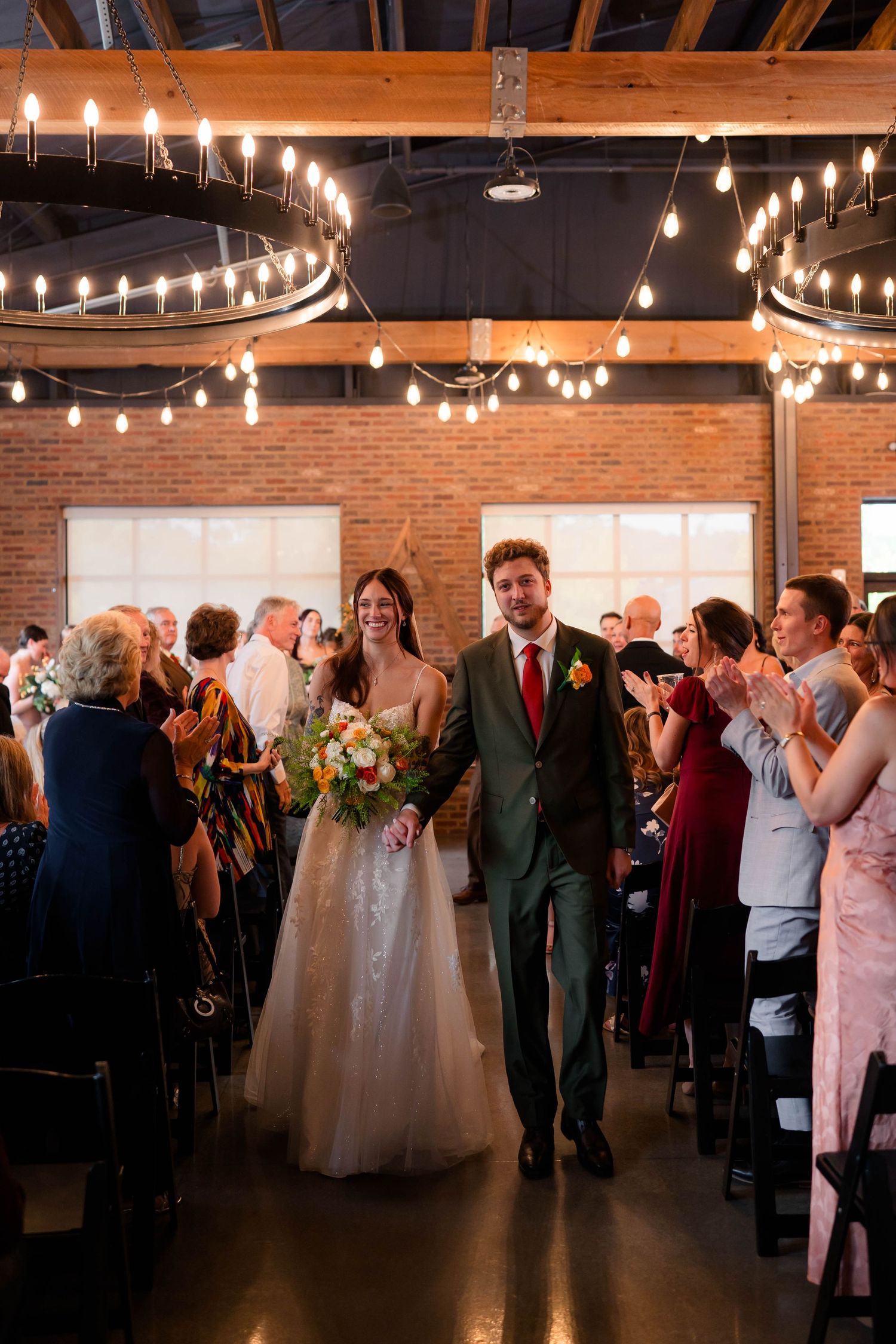 Newlyweds walk back down the aisle as guests applaud in warmly lit brick wedding venue with string lights.