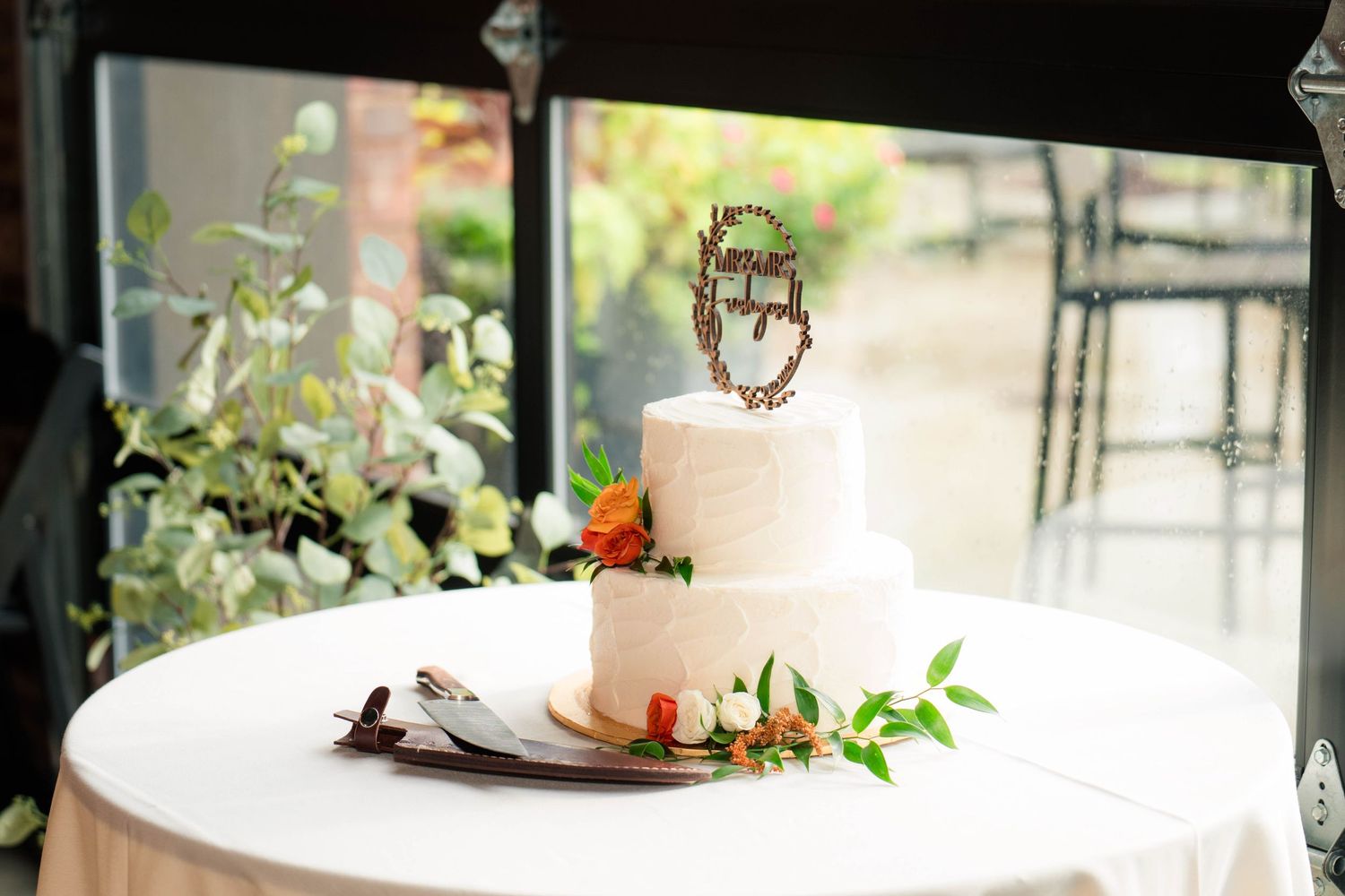 Two-tier white wedding cake with monogram topper and orange floral accents on draped table.