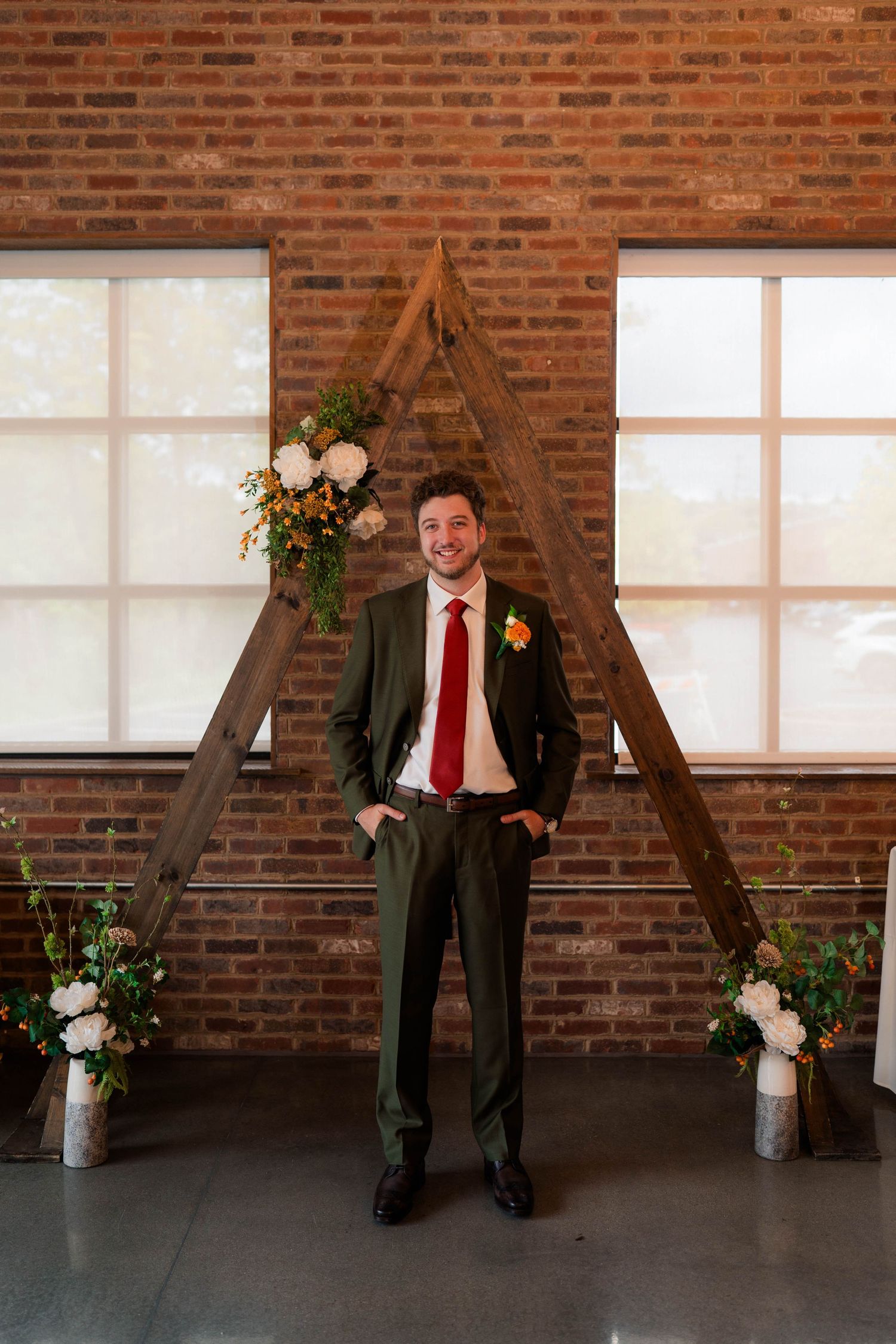 Groom in olive suit and red tie standing before a wooden triangular arch with floral arrangements.