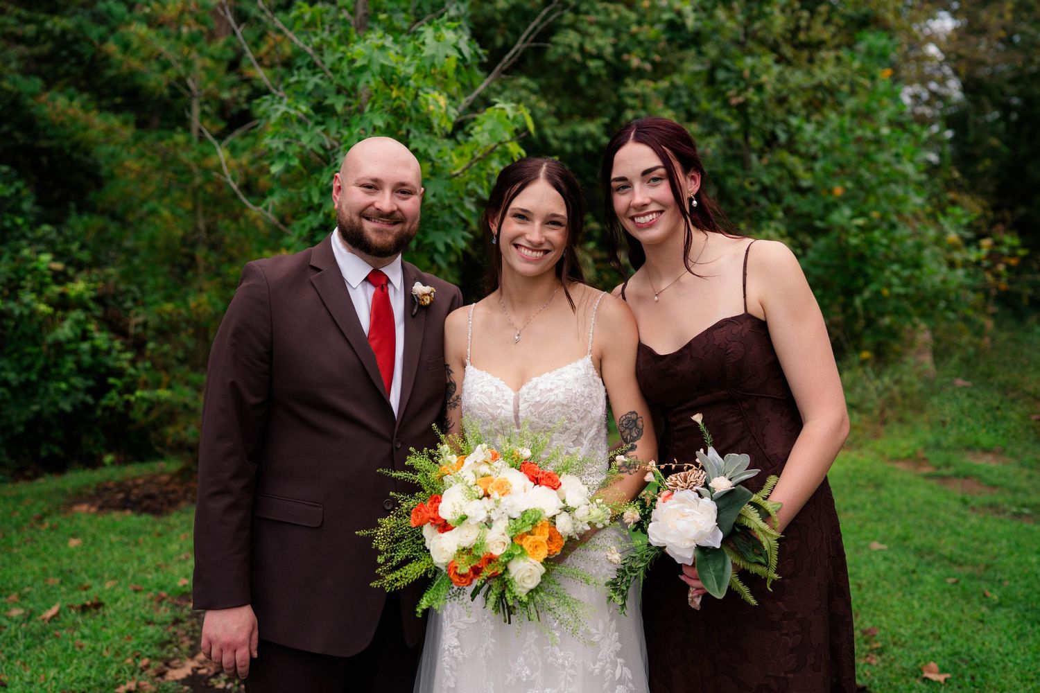 Bride with orange bouquet posing with man in brown suit and woman in dark bridesmaid dress.