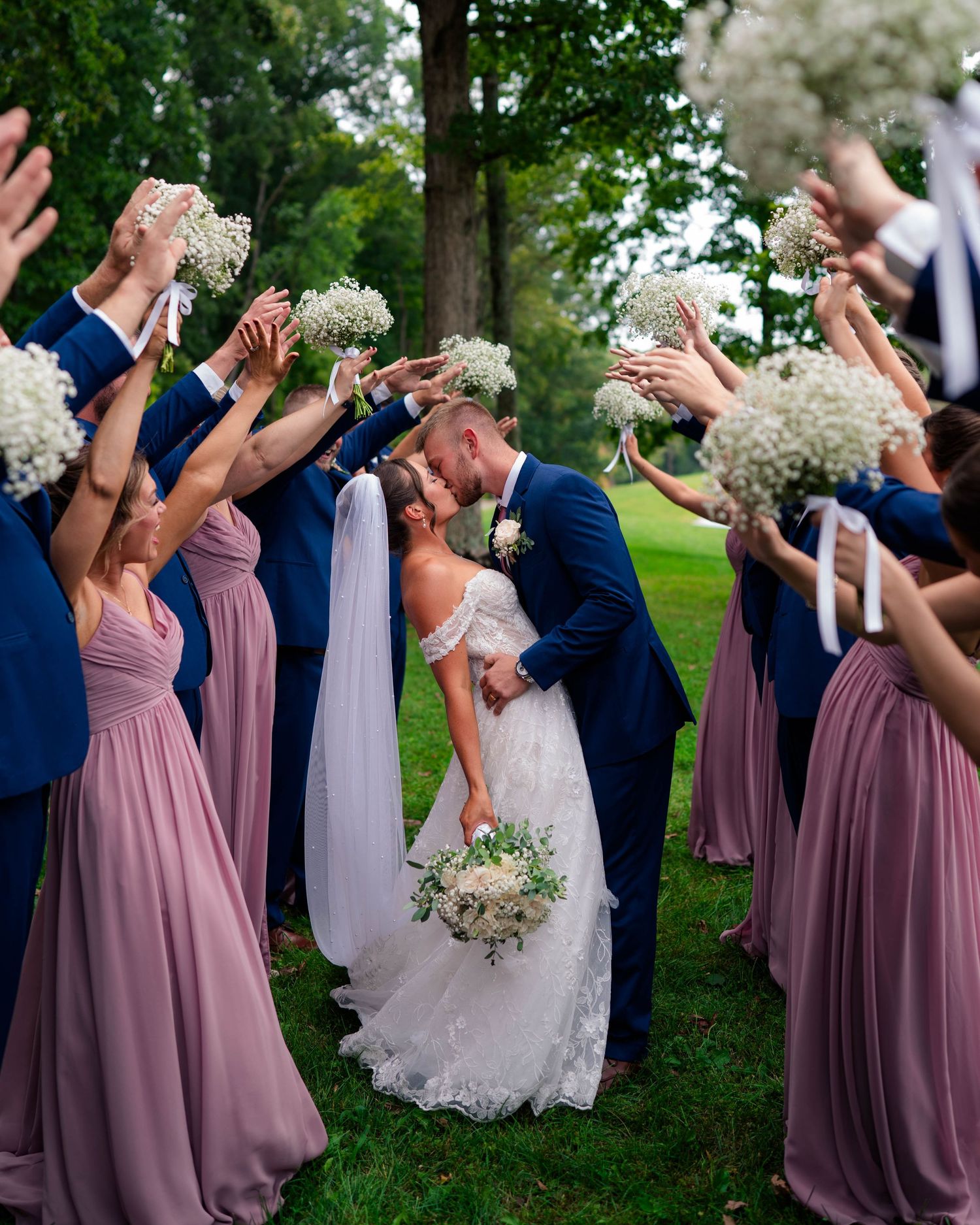 Bride and groom share a kiss under an arch of baby's breath bouquets held by bridesmaids and groomsmen.