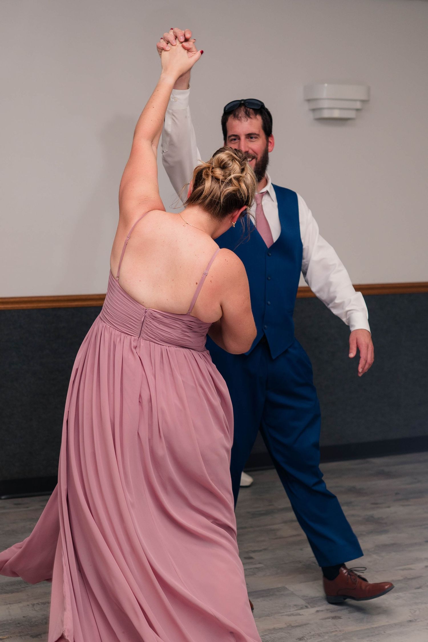 Bridesmaid in dusty rose gown dancing with bearded groomsman in navy suit at reception.