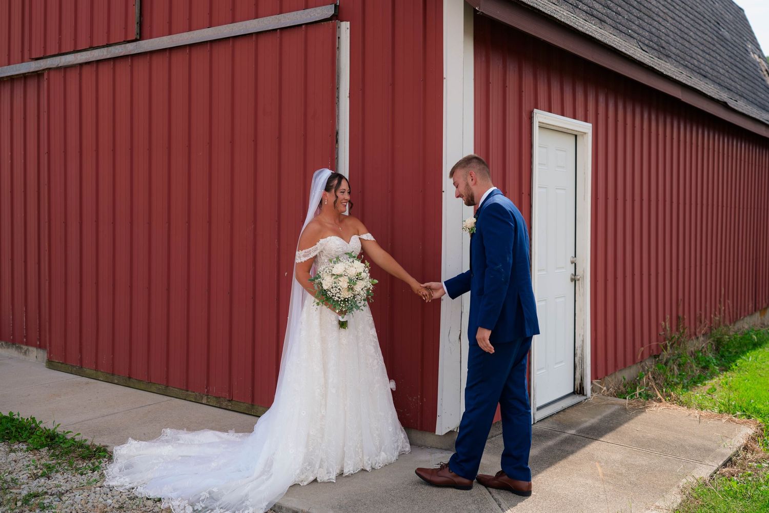 Bride and groom hold hands during first look outside a rustic red barn on their wedding day.