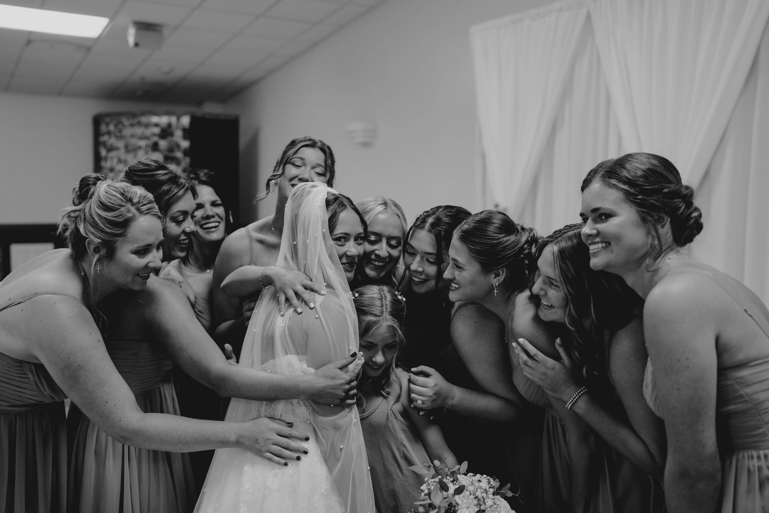 Black and white photo of bridesmaids surrounding and embracing bride in emotional wedding moment.