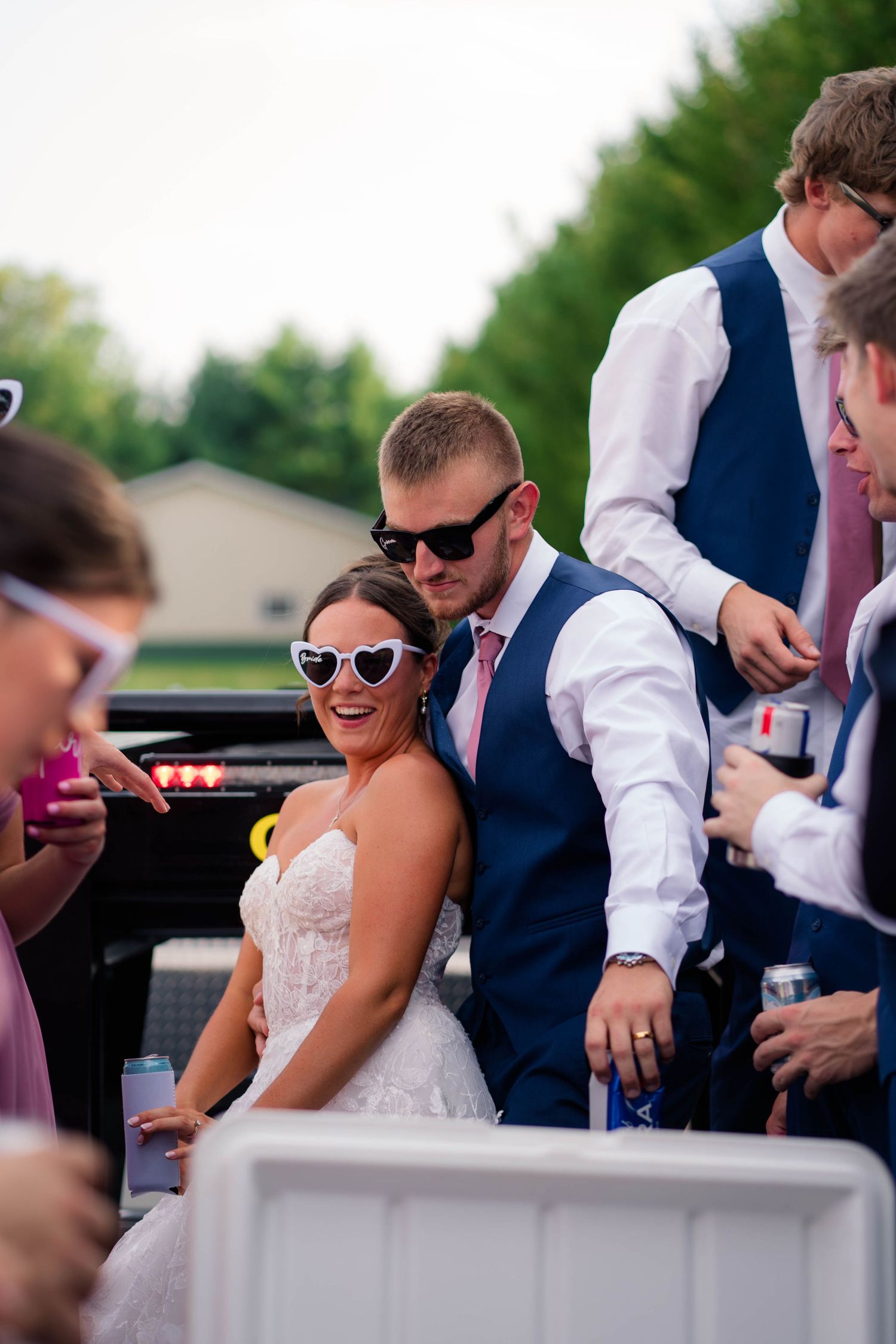 Bride and groom wearing heart sunglasses laughing with wedding guests in truck bed at outdoor reception.