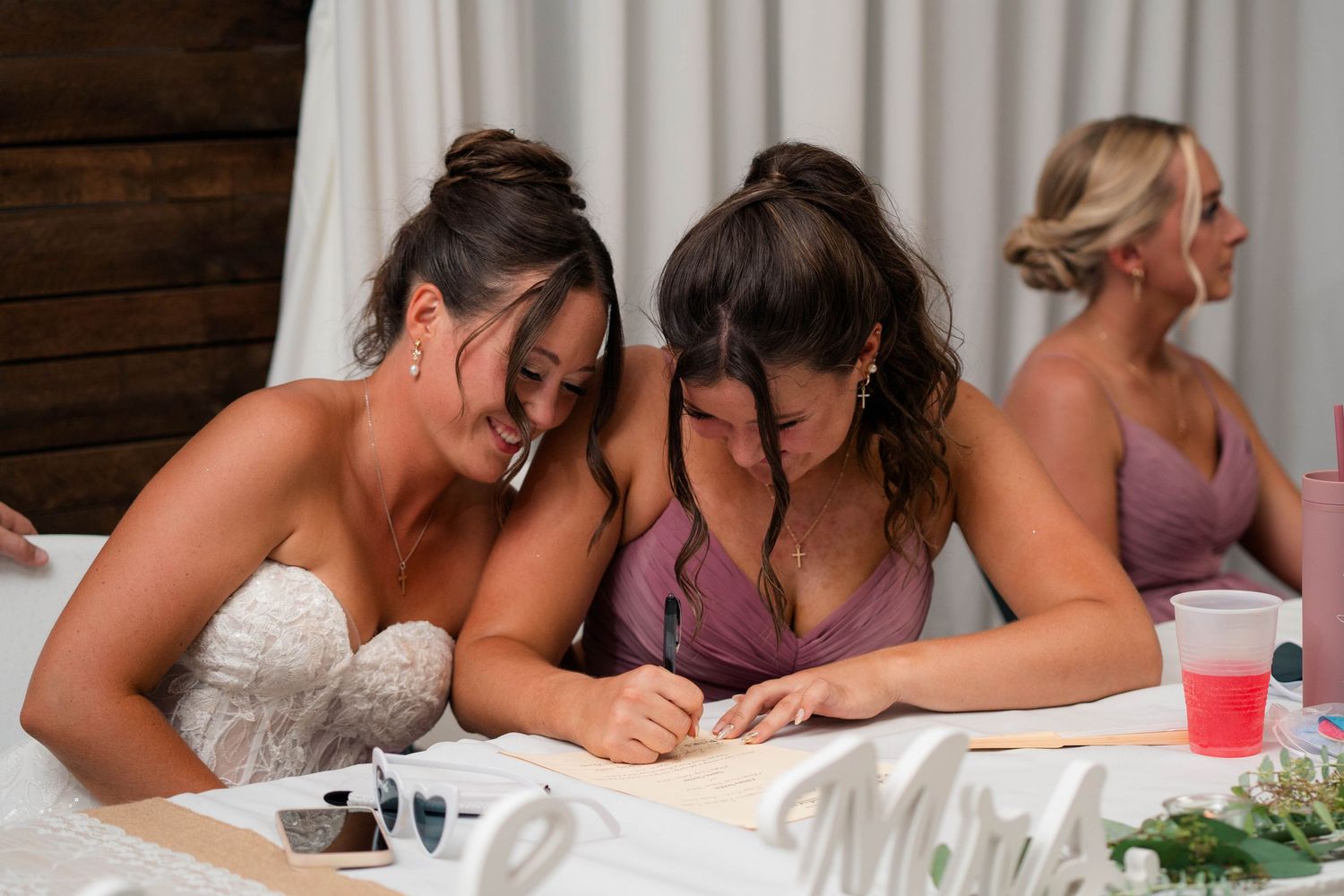 Bride and bridesmaid laugh together while signing documents at wedding reception table.