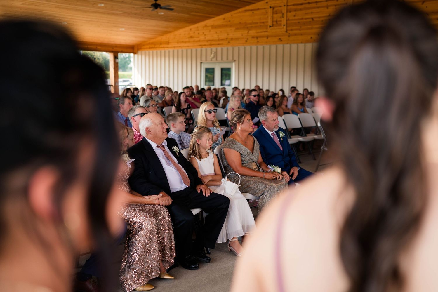 Wedding guests seated under pavilion watch outdoor ceremony from audience perspective through bridal party foreground.