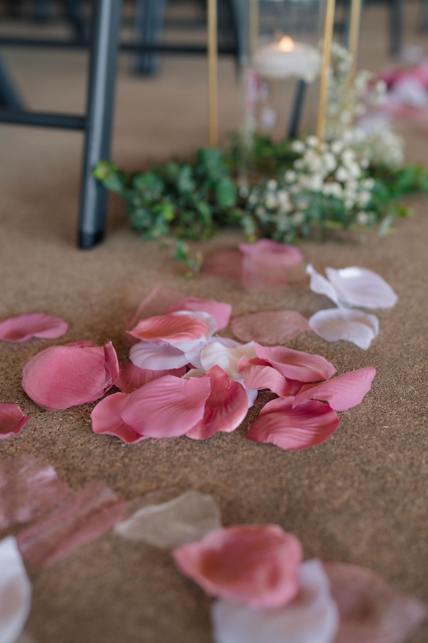Pink and white rose petals scattered on sandy floor with floral centerpiece and candle in background at wedding.