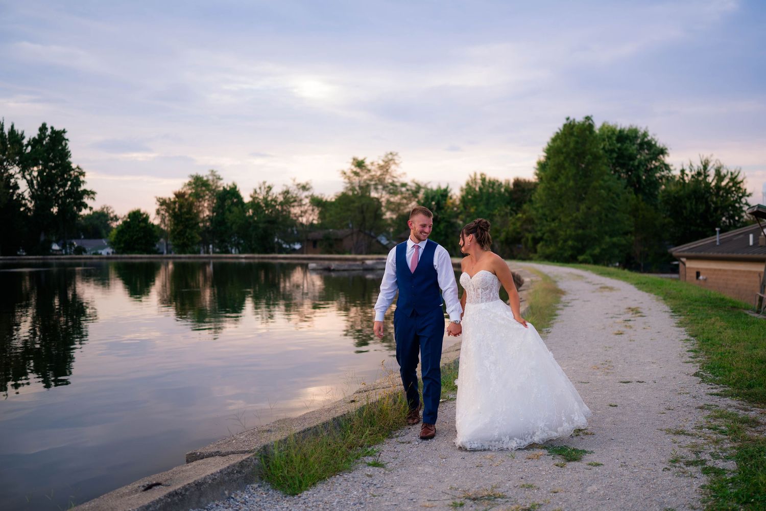 Bride and groom walk hand in hand along a scenic lakeside path at sunset on their wedding day.
