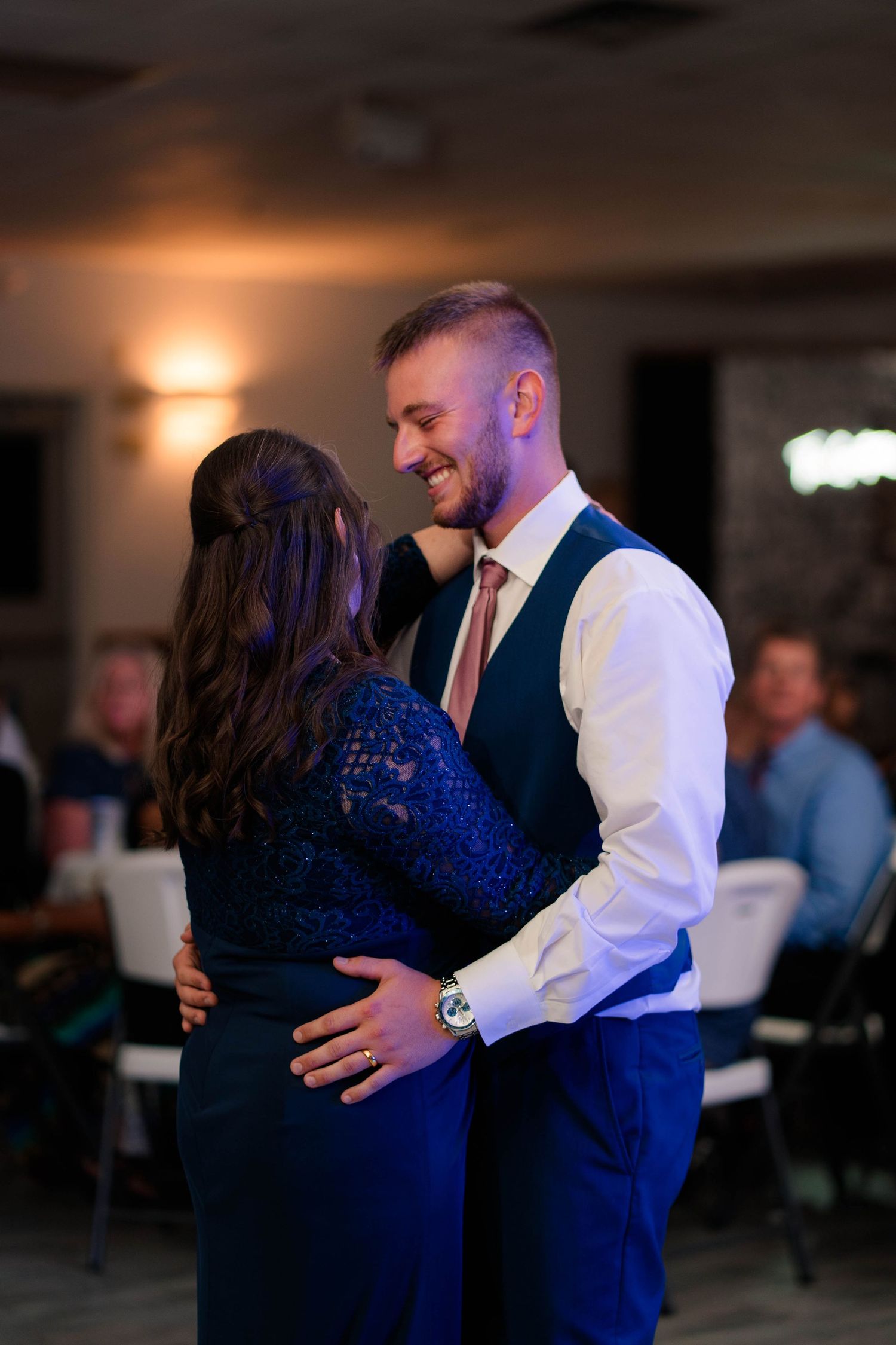 Groom in navy vest smiles while sharing mother-son dance at wedding reception.