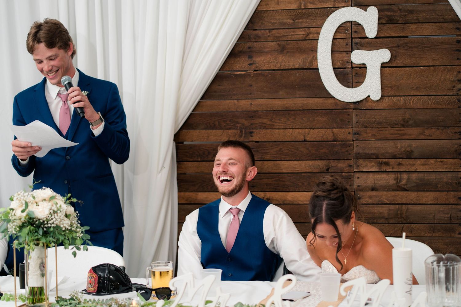 Best man reads a humorous speech as the groom laughs and bride covers her face at an elegant wedding reception.