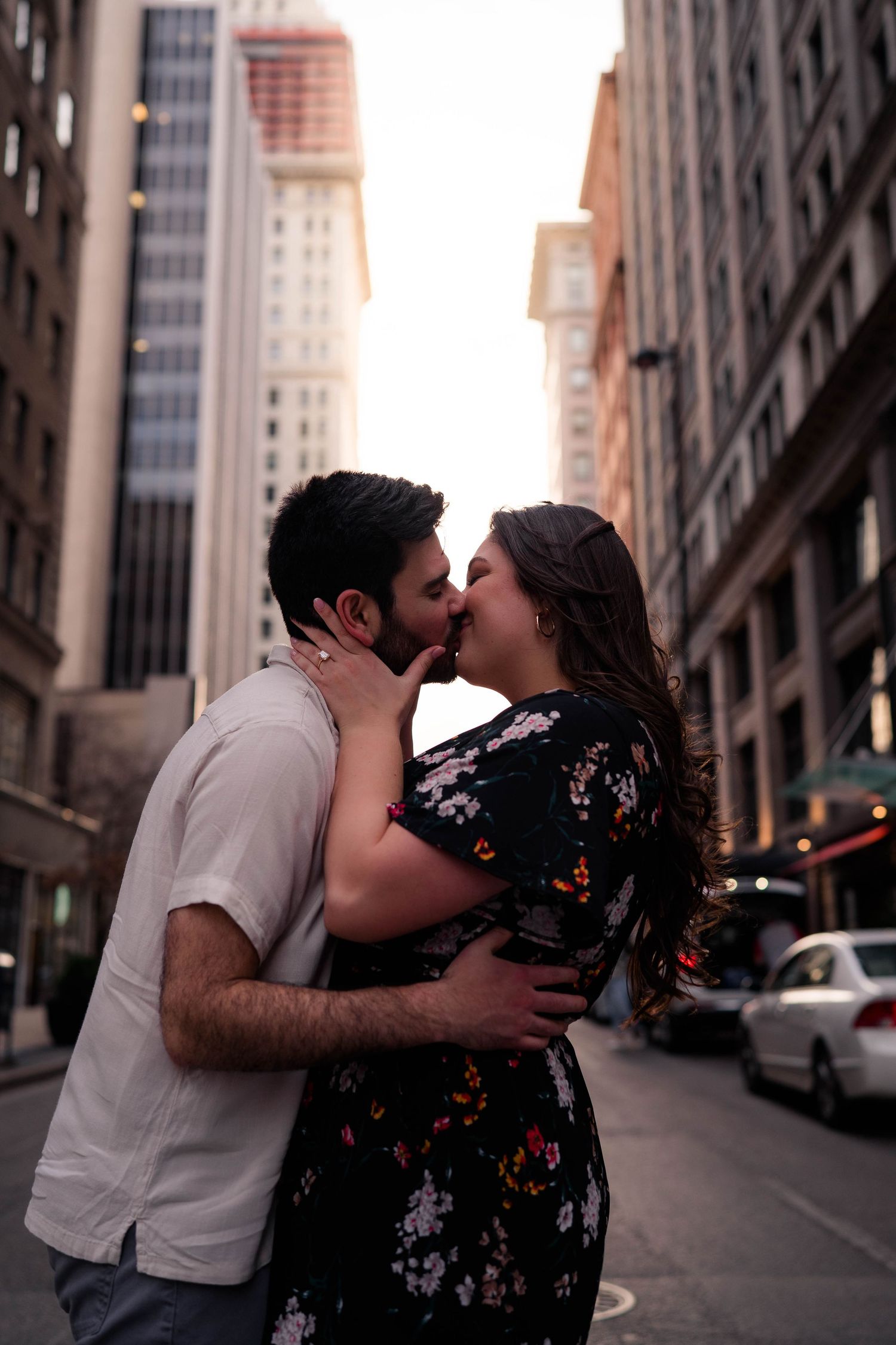 Couple sharing a romantic kiss on a city street surrounded by tall buildings at sunset.