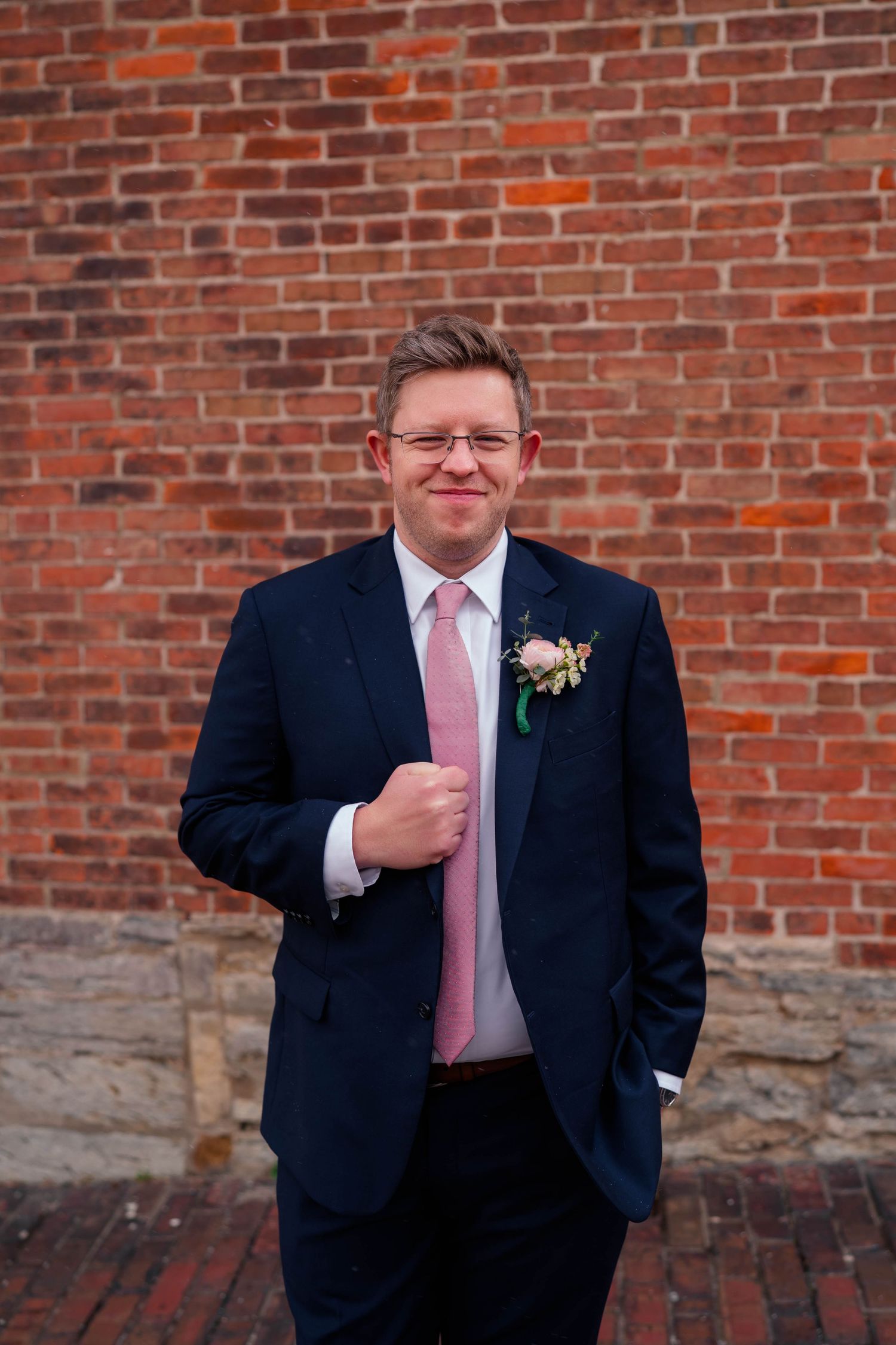 A groom in a navy suit and pink tie with a boutonniere smiles in front of a red brick wall.