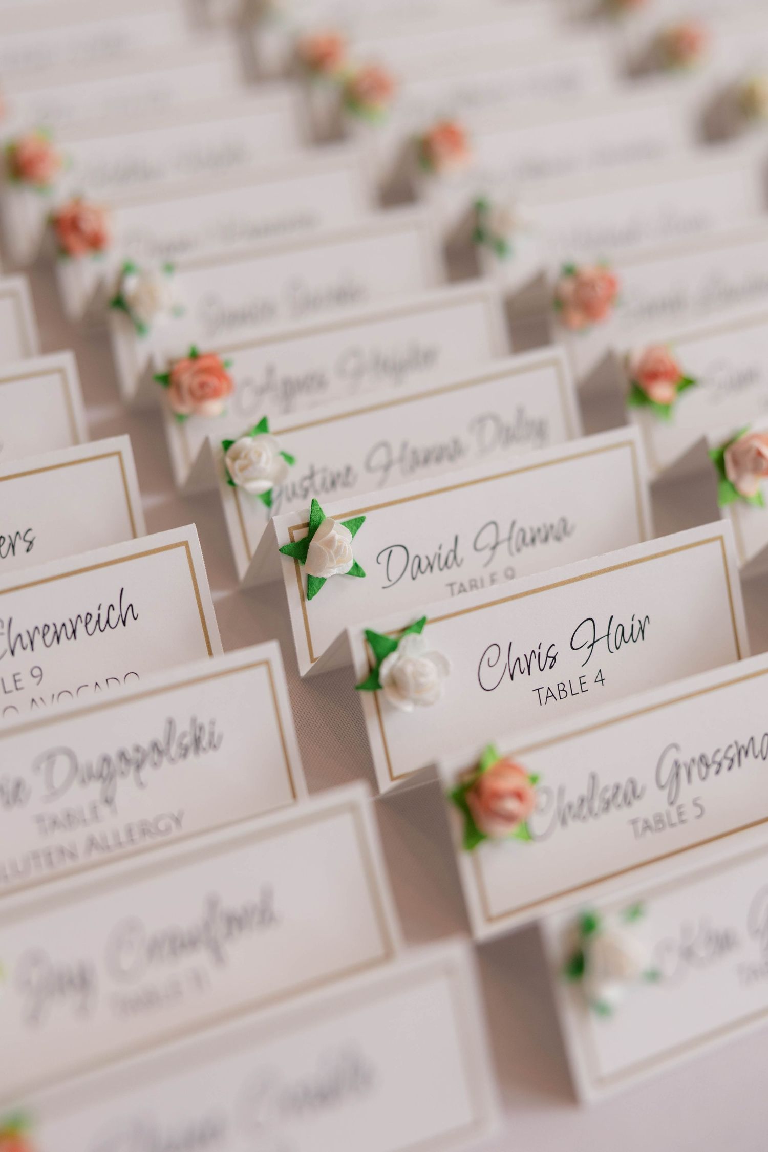 Close-up of elegant wedding place cards decorated with pink and white roses, displaying guest names and table numbers in script.