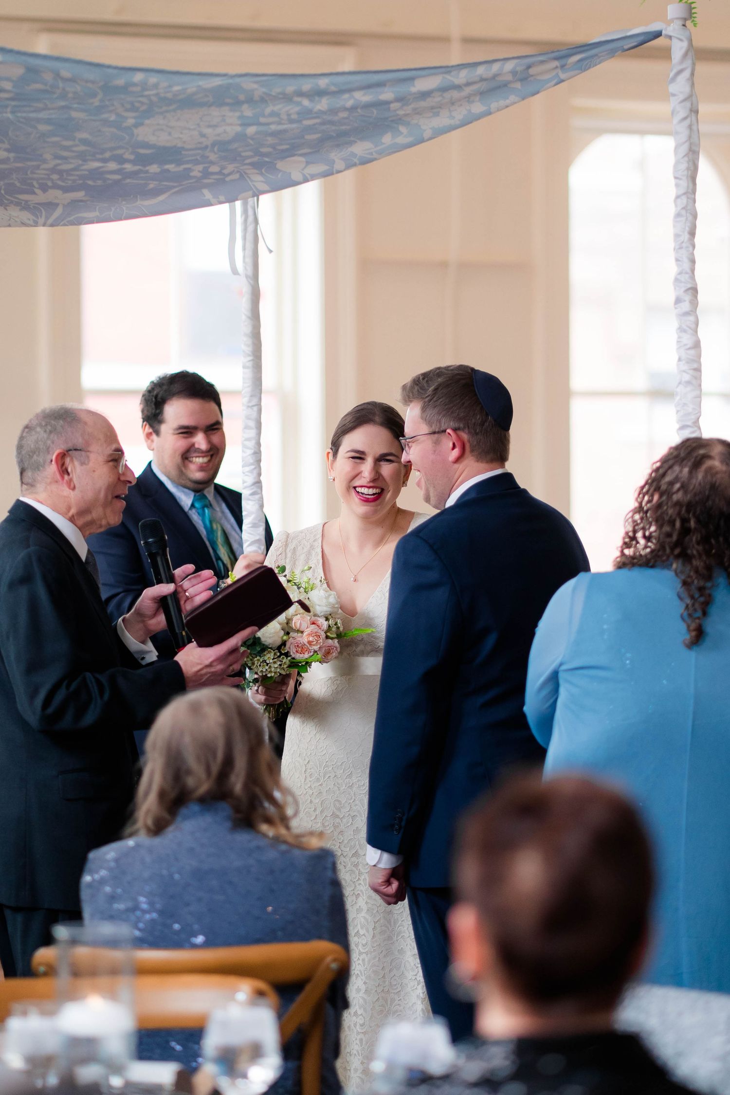 Bride laughs joyfully with groom and officiant during Jewish wedding ceremony under chuppah.
