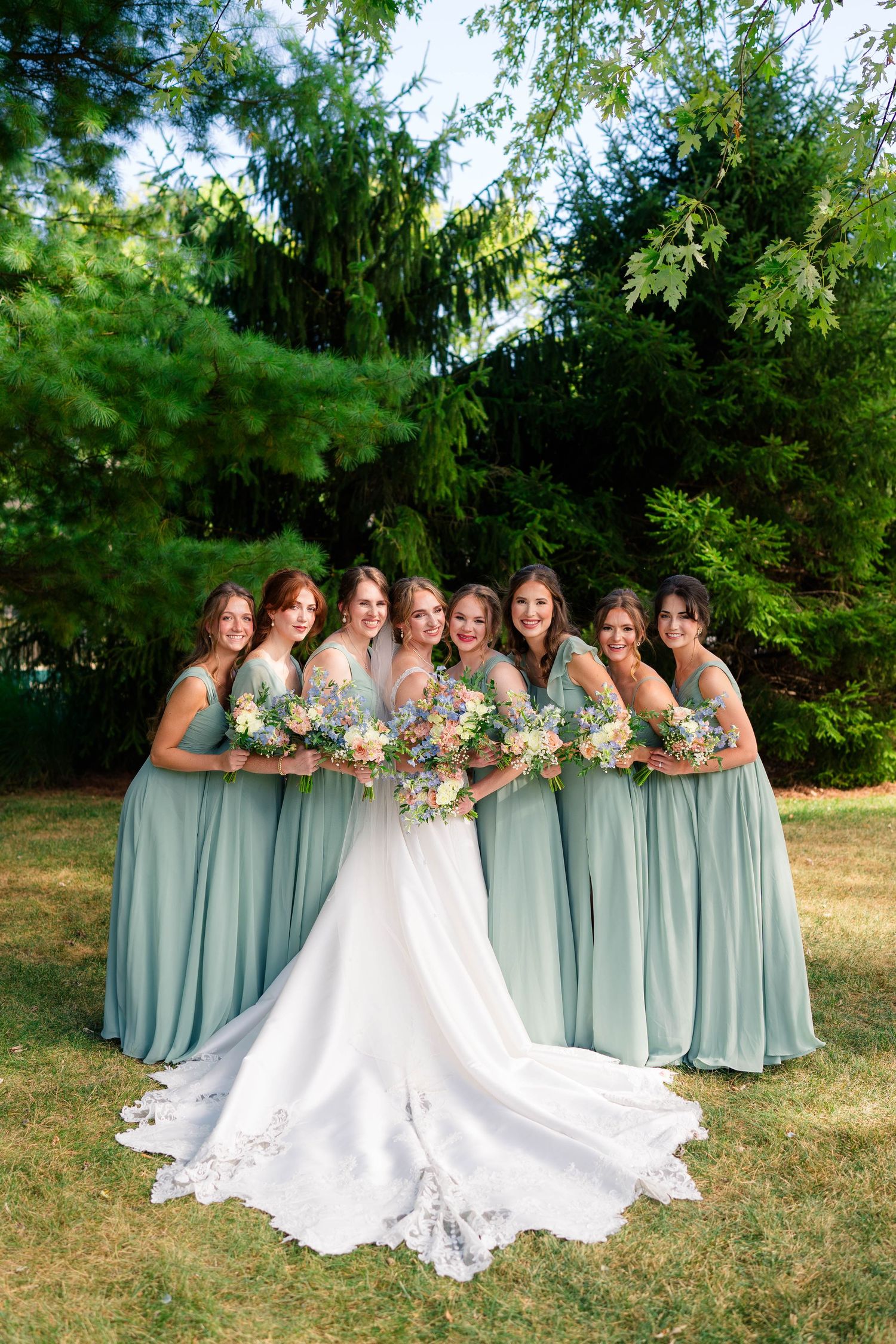 Bride with bridesmaids in sage green gowns holding bouquets under lush green trees outdoors.