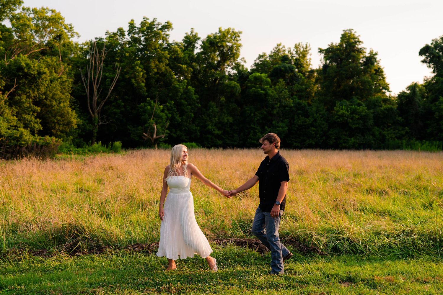 A couple holds hands in a golden meadow at sunset, surrounded by lush green trees, the woman in white dress.