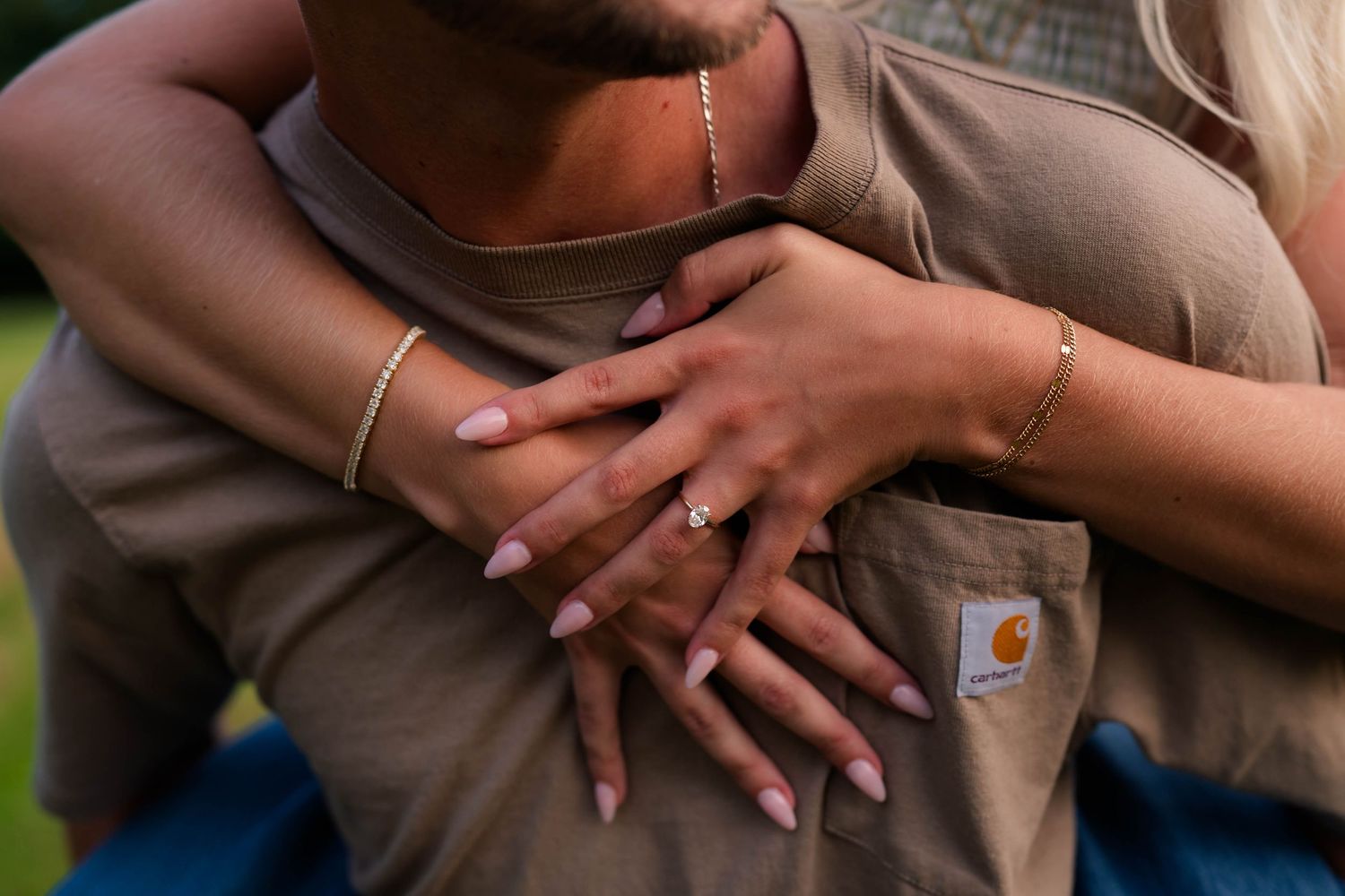 Close-up of couple embracing, woman showing engagement ring with gold bracelets, man wearing Carhartt shirt outdoors.