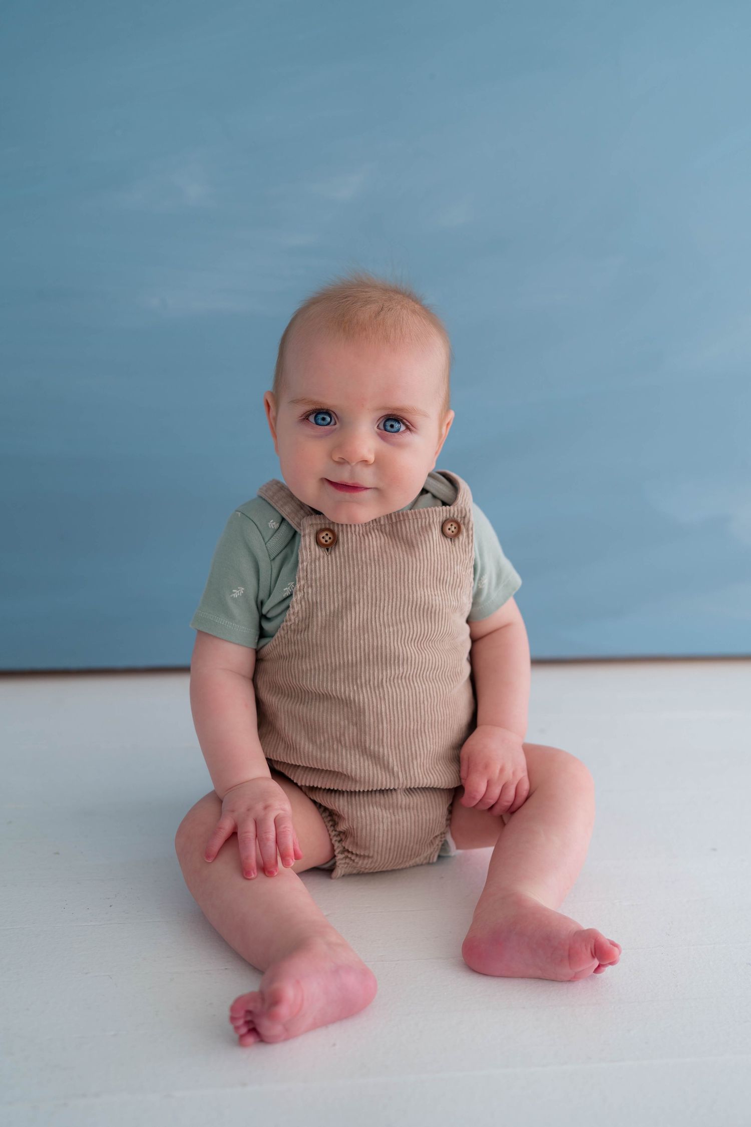 Adorable blue-eyed baby sitting upright in a tan corduroy romper against a blue background.