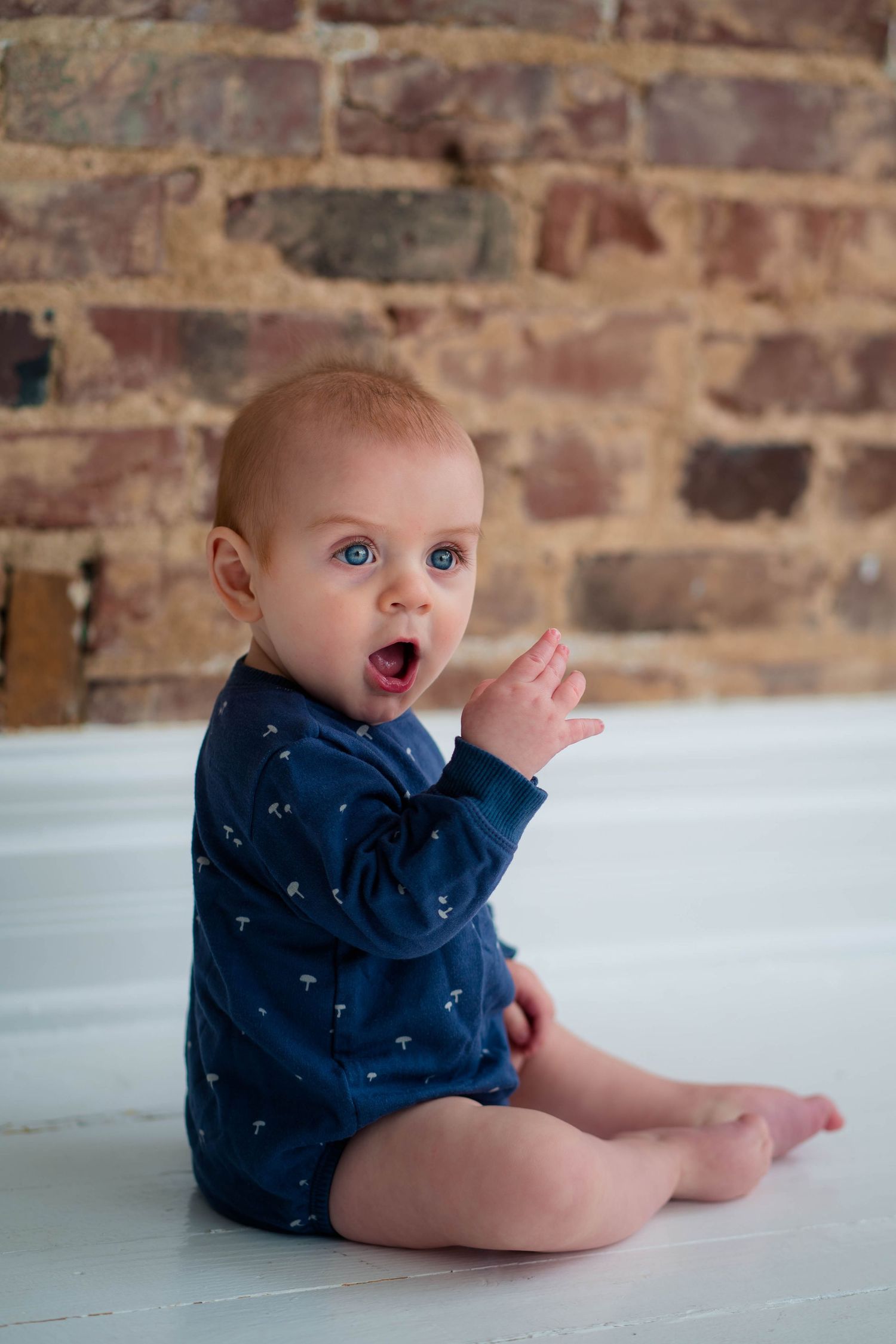 Adorable blue-eyed baby in navy bodysuit sitting on white floor near brick wall.