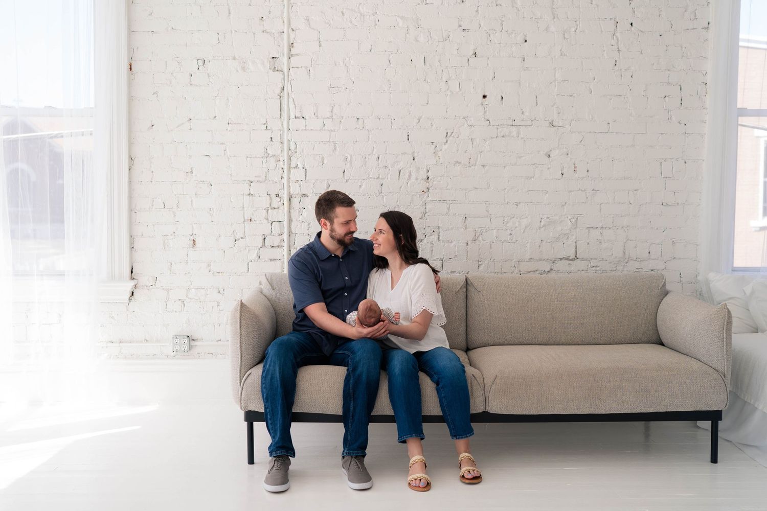 Loving couple gazing at each other on a couch while holding their newborn in a white brick studio.