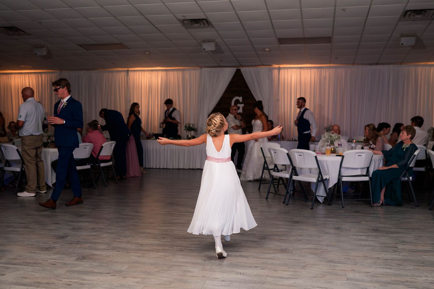 Young flower girl twirls gracefully on the dance floor during a wedding reception in a decorated banquet hall.