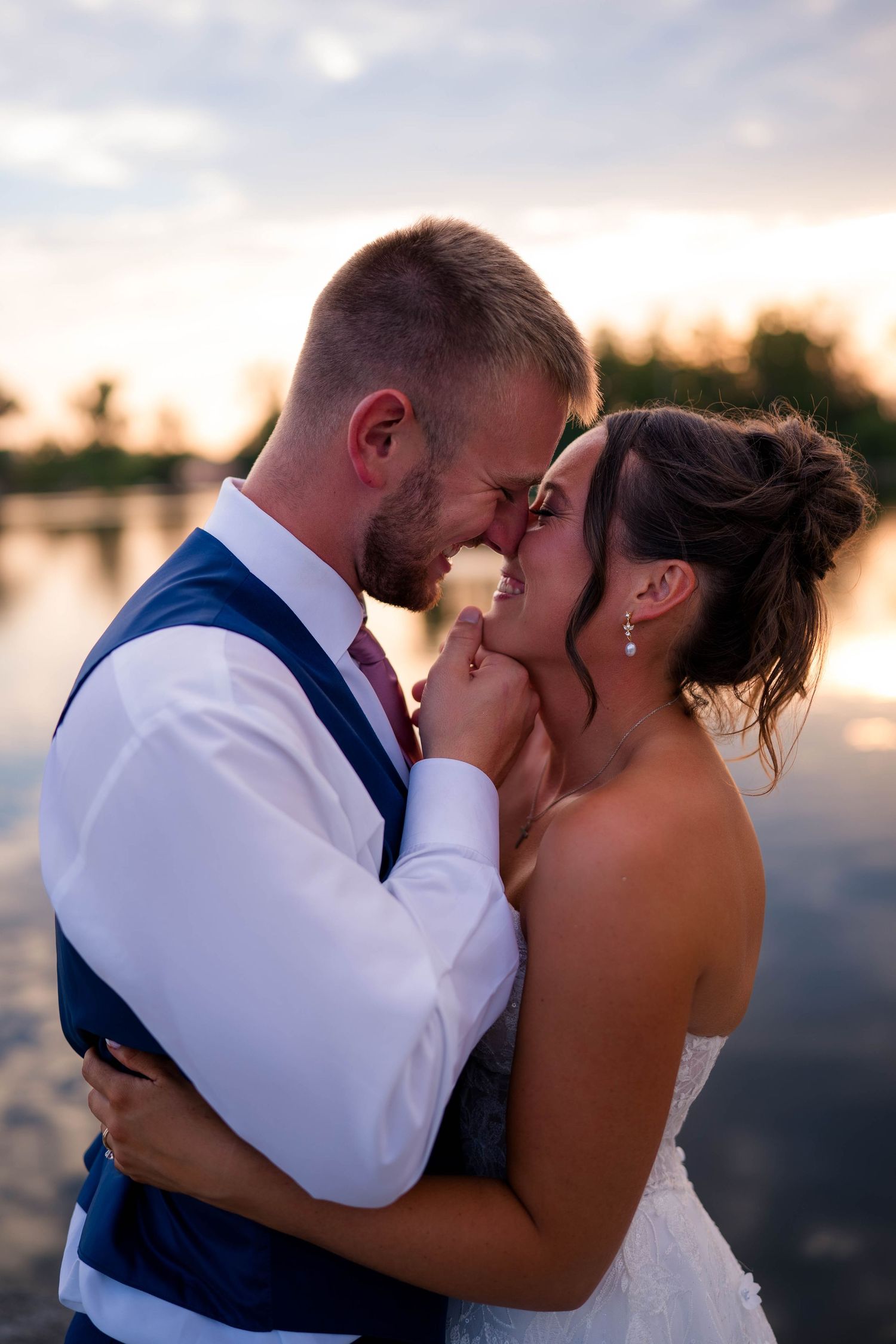Bride and groom share a romantic moment by a lake at sunset, with the groom tenderly holding the bride's face.