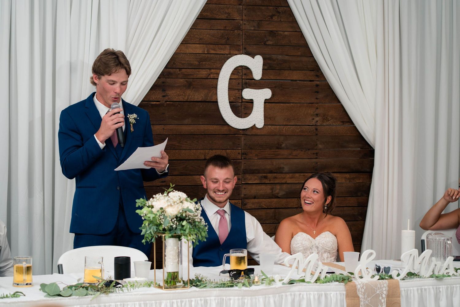Man in blue suit gives wedding toast as bride and groom smile at decorated head table with floral arrangements.