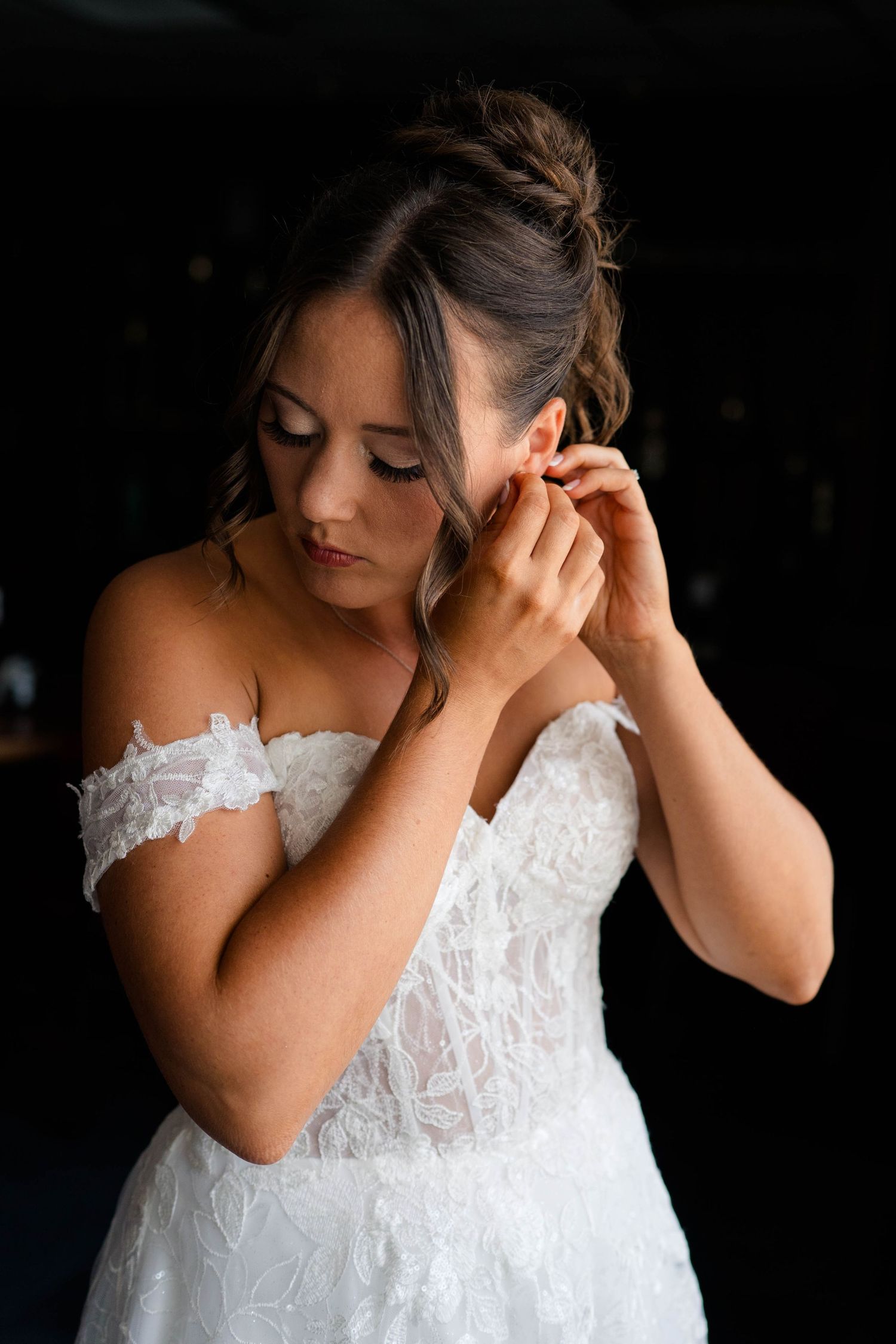 Bride in off-shoulder lace gown putting on earring while getting ready on her wedding day.