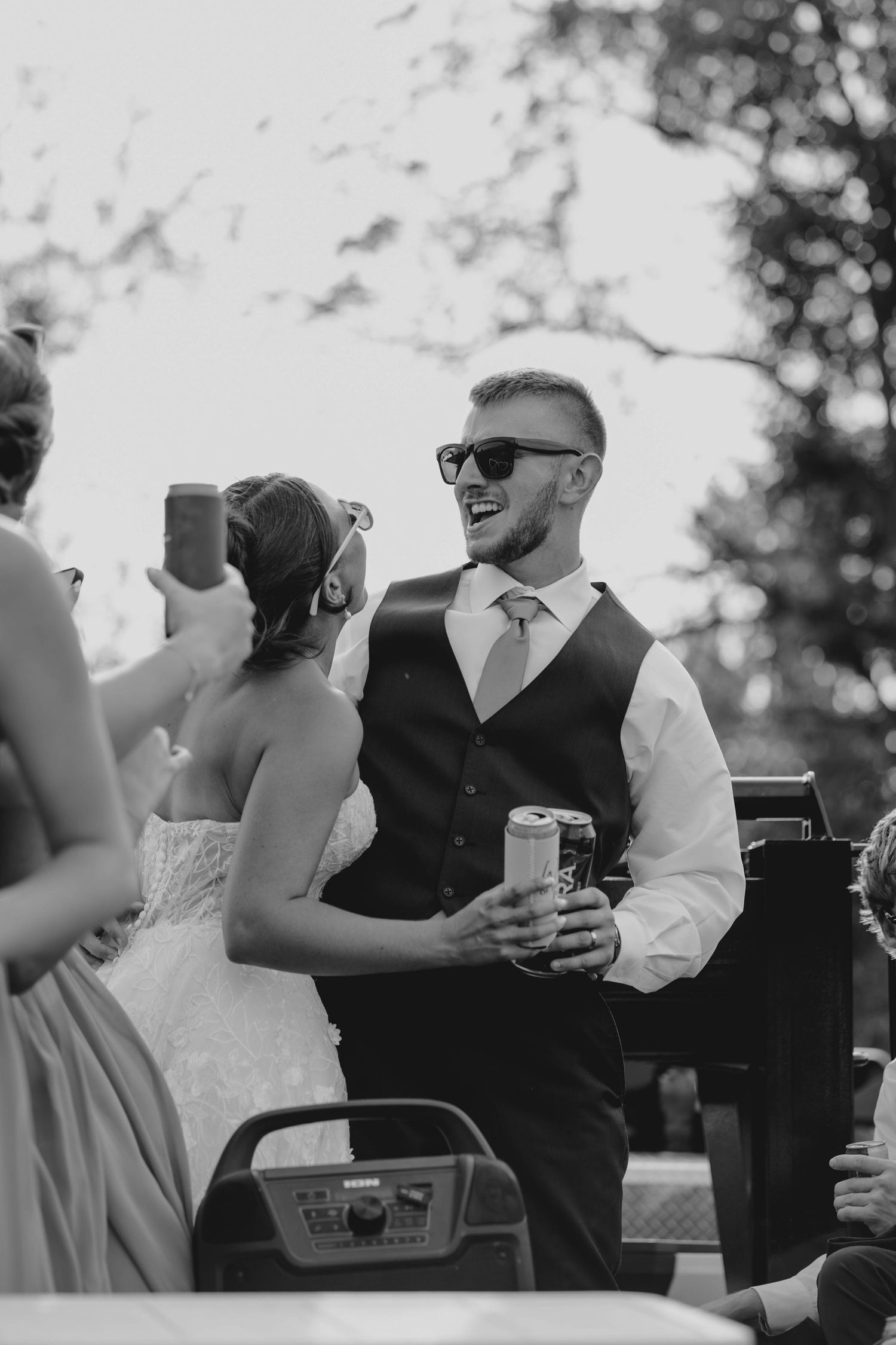 Black and white photo of laughing groom in sunglasses holding beer can at wedding reception.