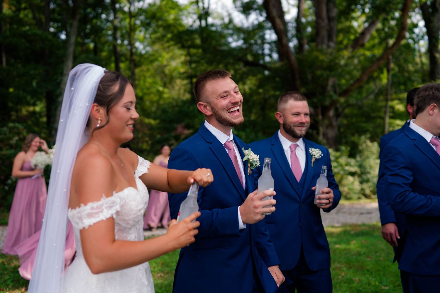 Laughing bride and groom celebrate outdoors with wedding party members in pink and navy.