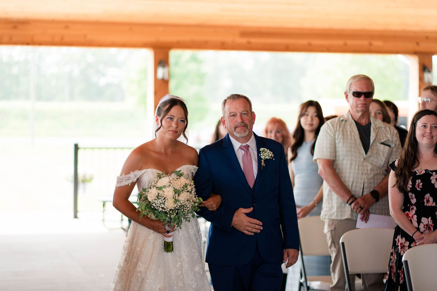 Bride in off-shoulder lace gown walks down the aisle with escort during outdoor wedding ceremony.