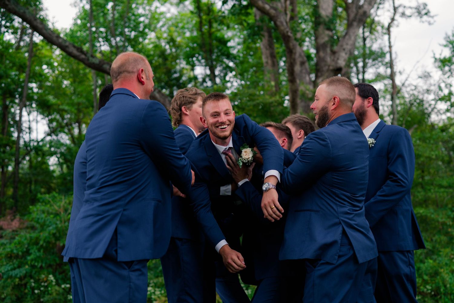 Groomsmen in navy suits laughing and celebrating together with groom in wooded outdoor setting.