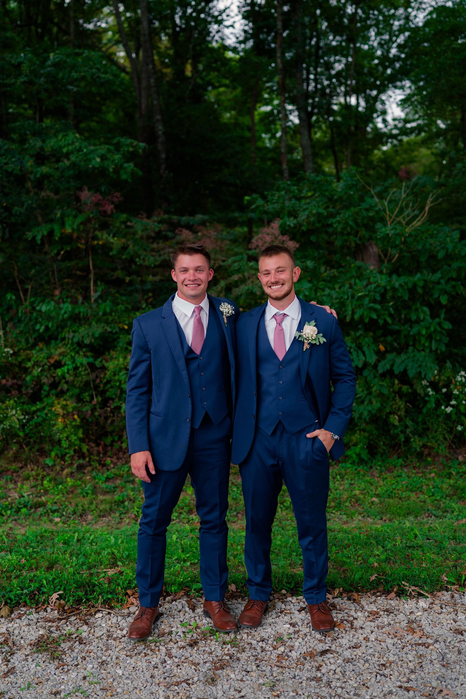 Two groomsmen in navy blue suits with pink ties posing outdoors at a wedding.