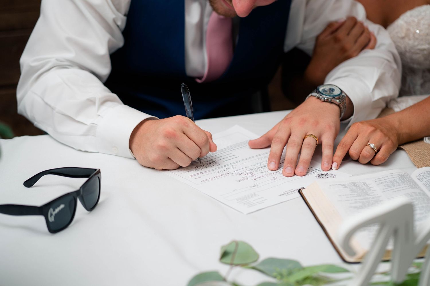 Groom signing wedding certificate with bride's hand resting on document beside sunglasses on table.