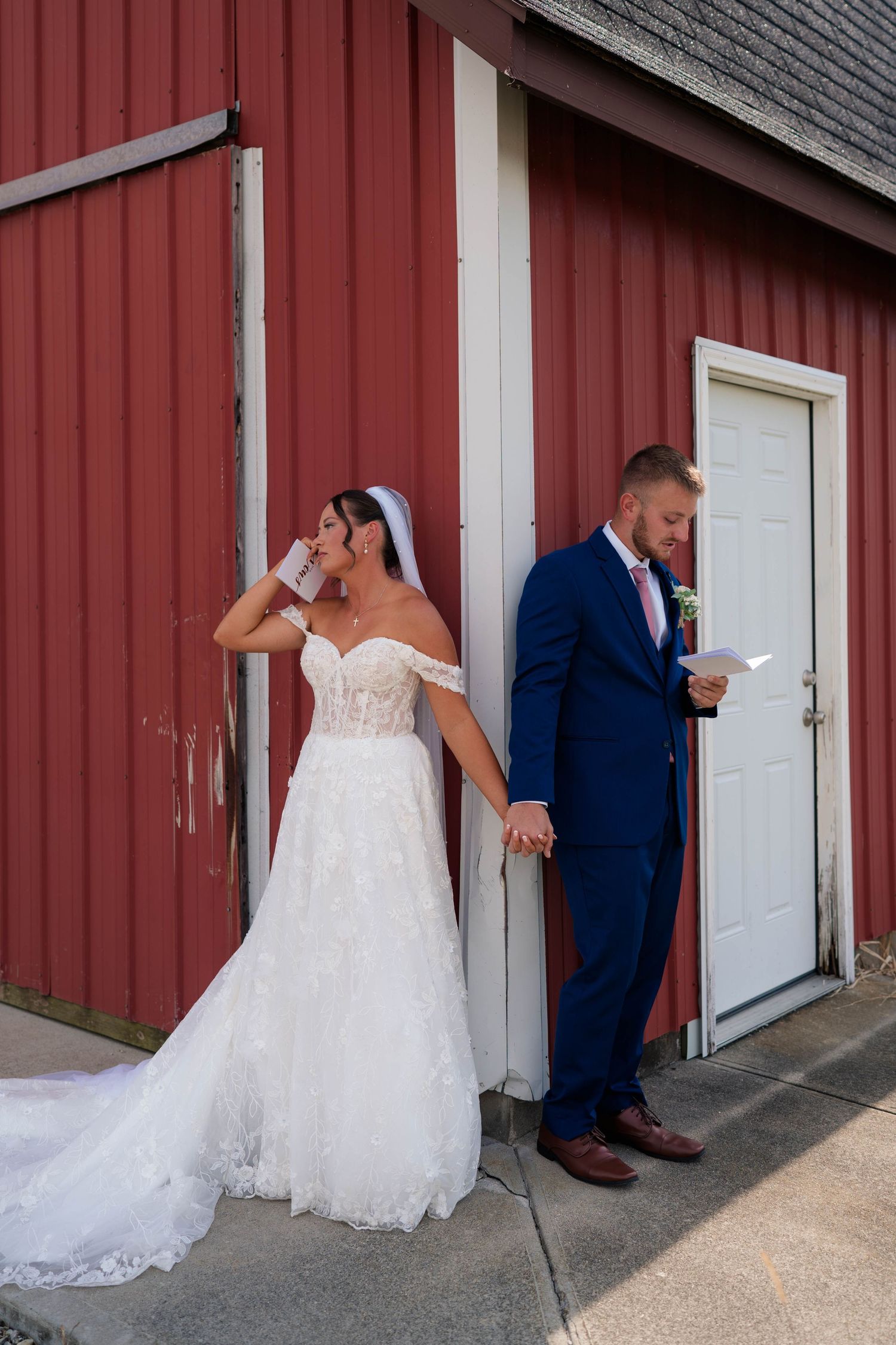 Emotional bride wipes tears while groom reads vows holding hands outside a red barn.