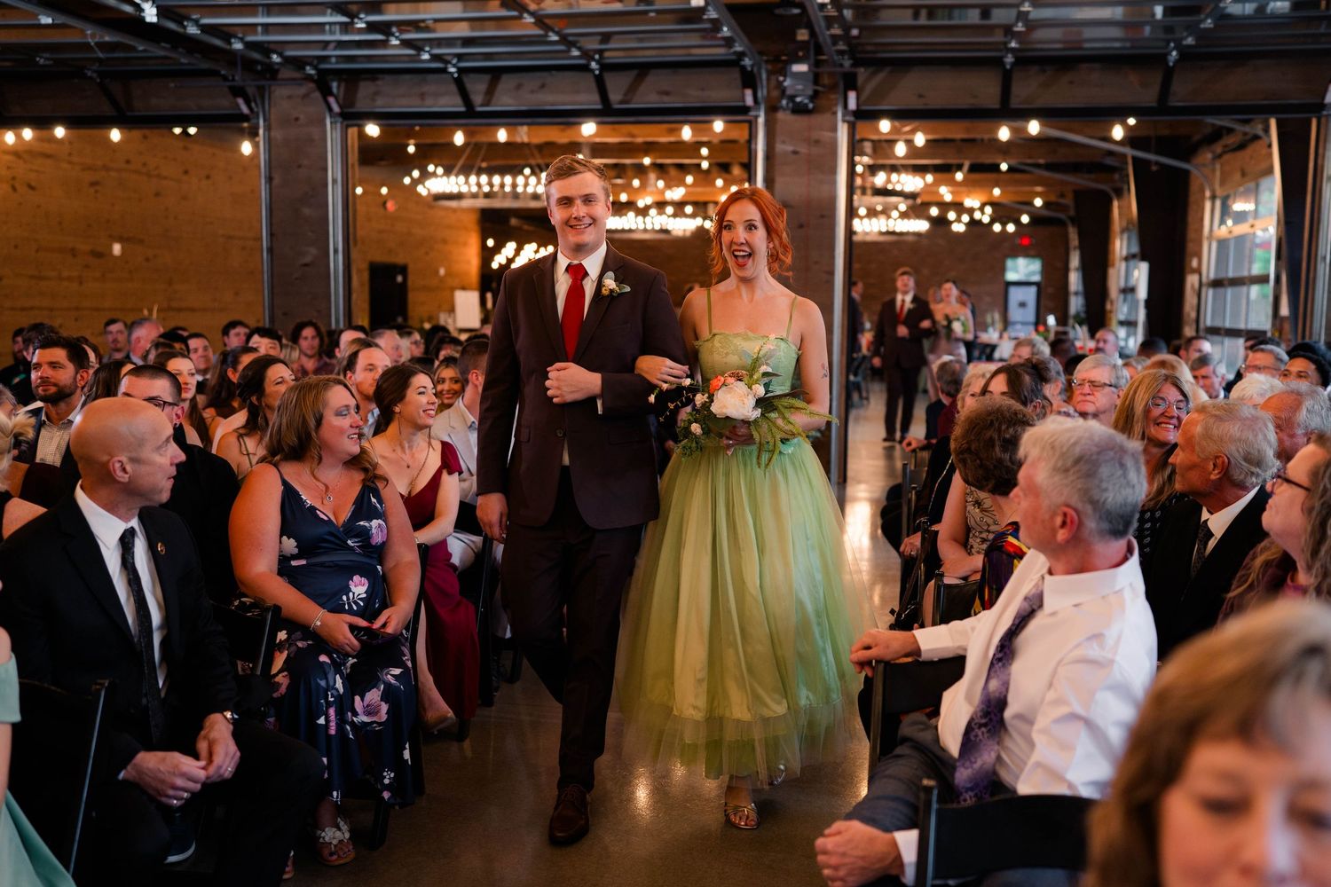 Groomsman escorting woman in green gown down wedding ceremony aisle before guests.