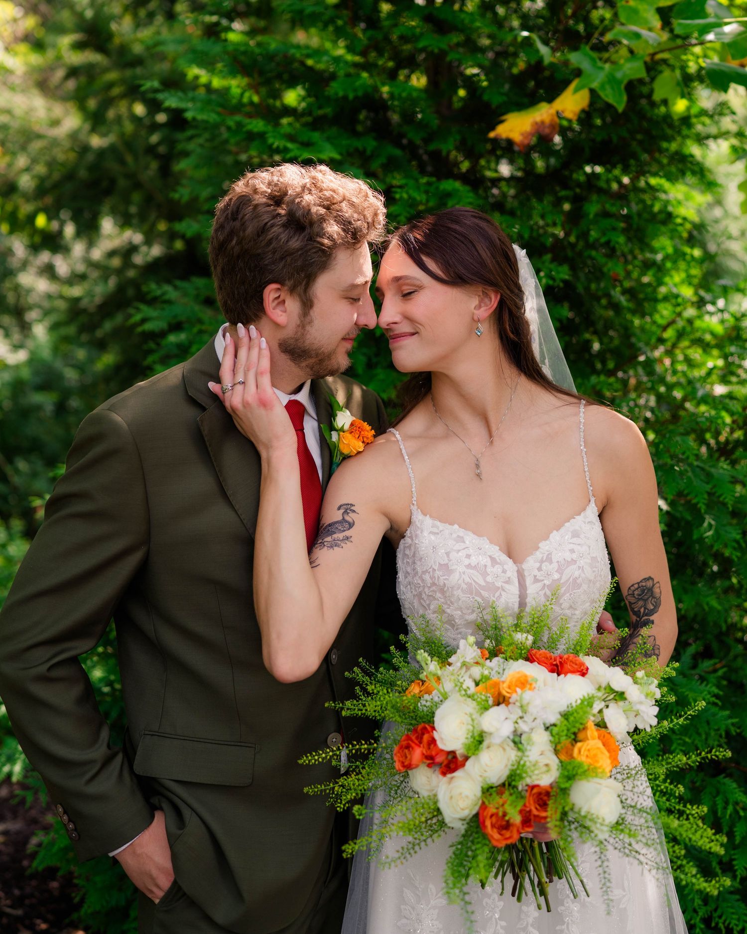 Bride and groom touch foreheads tenderly outdoors surrounded by lush green foliage, holding a vibrant bouquet.