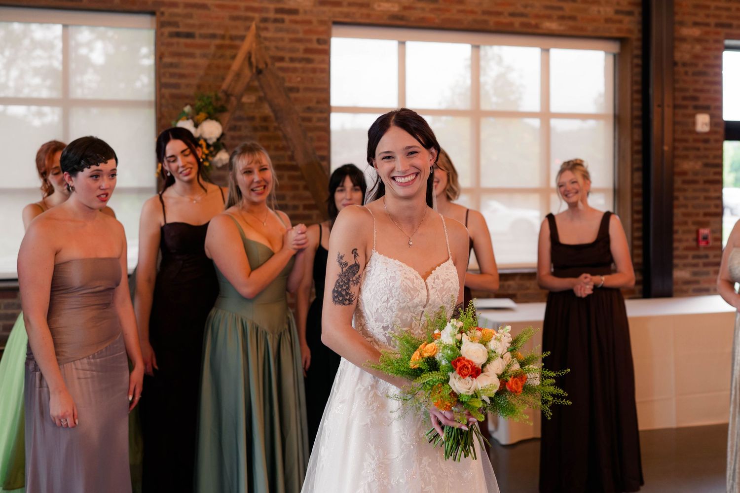Smiling bride in lace gown holds vibrant bouquet while bridesmaids in mixed earth-tone dresses stand behind her.