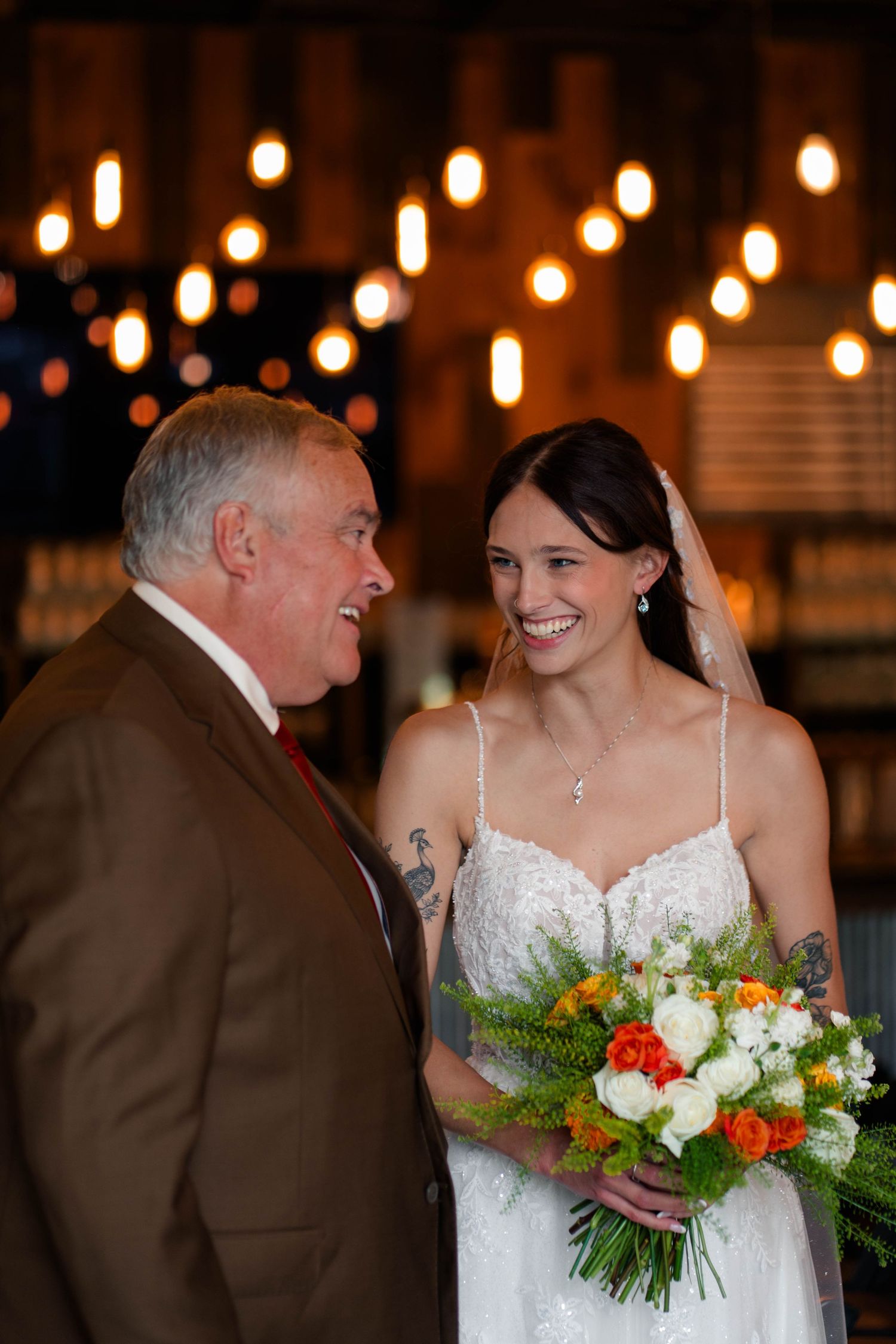 Smiling bride in lace gown holding colorful bouquet with older man in brown suit.