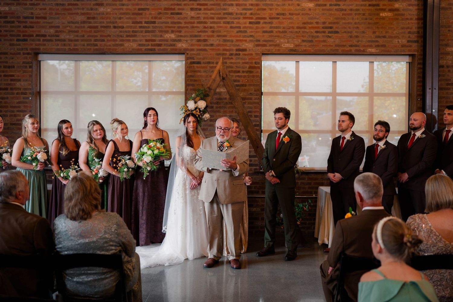 Wedding party stands at altar in rustic brick venue with bridesmaids in purple dresses and groomsmen in green suits.