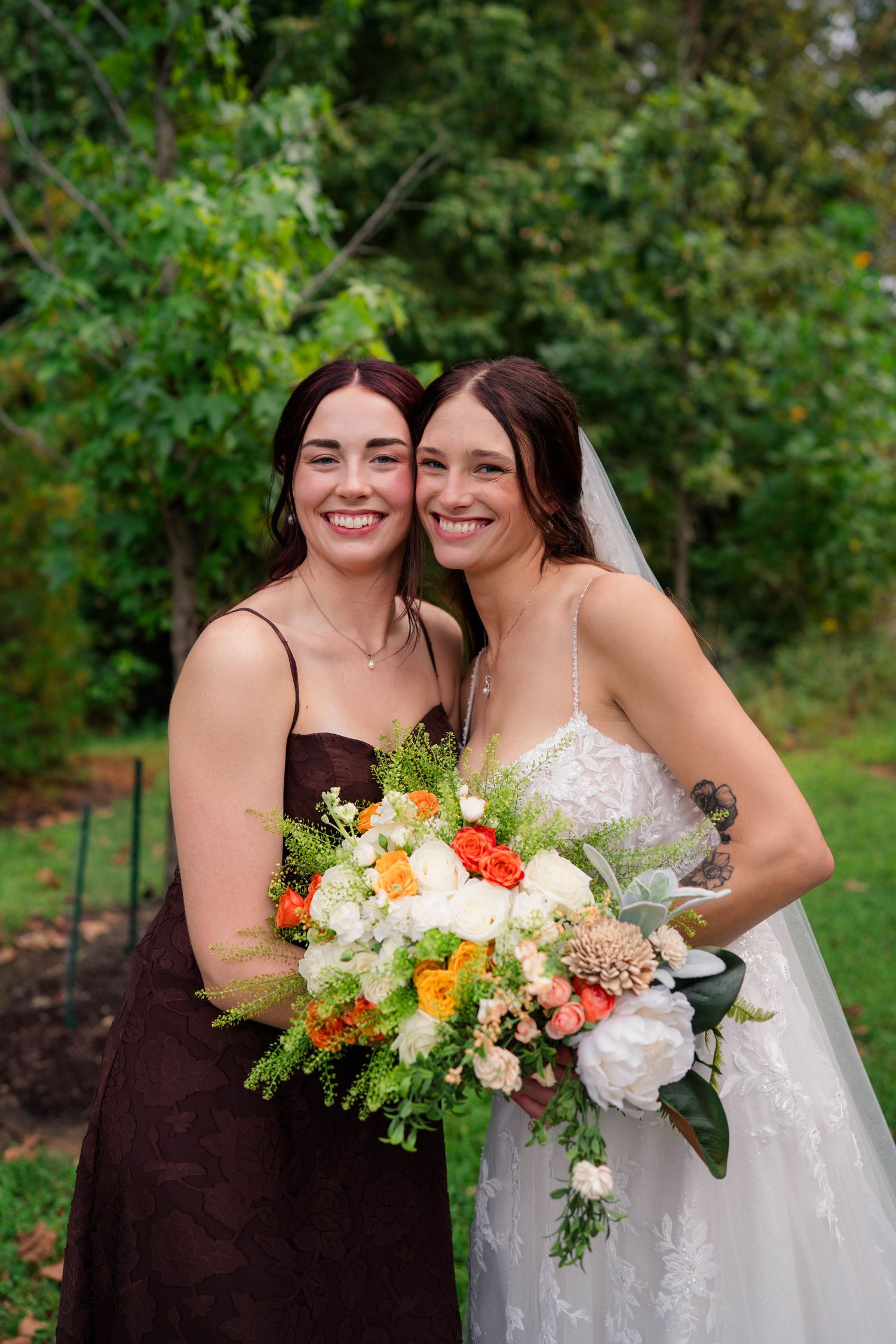 Bride in lace gown and bridesmaid in brown dress smile together holding colorful wedding bouquets.
