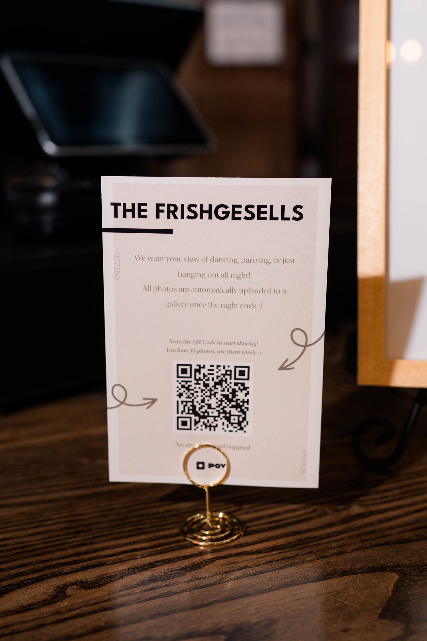 The Frishgesells wedding QR code photo sharing sign displayed in gold holder on wood table.