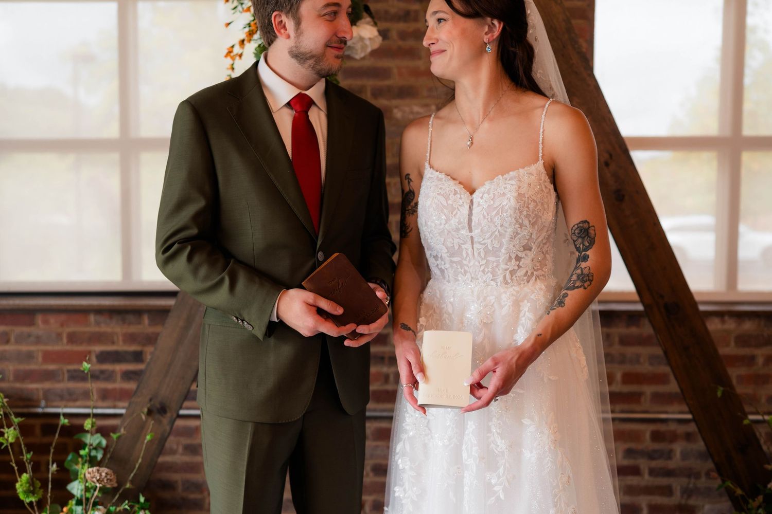 Bride and groom exchange vows holding books at rustic brick wall wedding ceremony venue.
