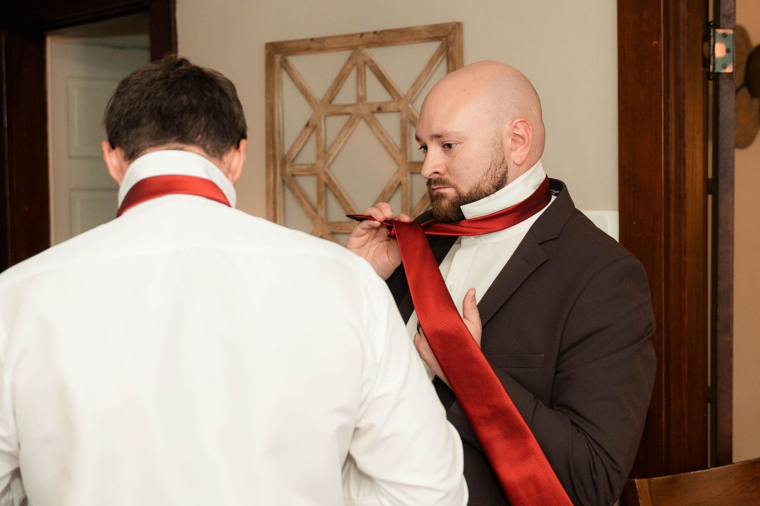 A bald groom adjusts a red tie on a groomsman in a white shirt before a wedding ceremony.