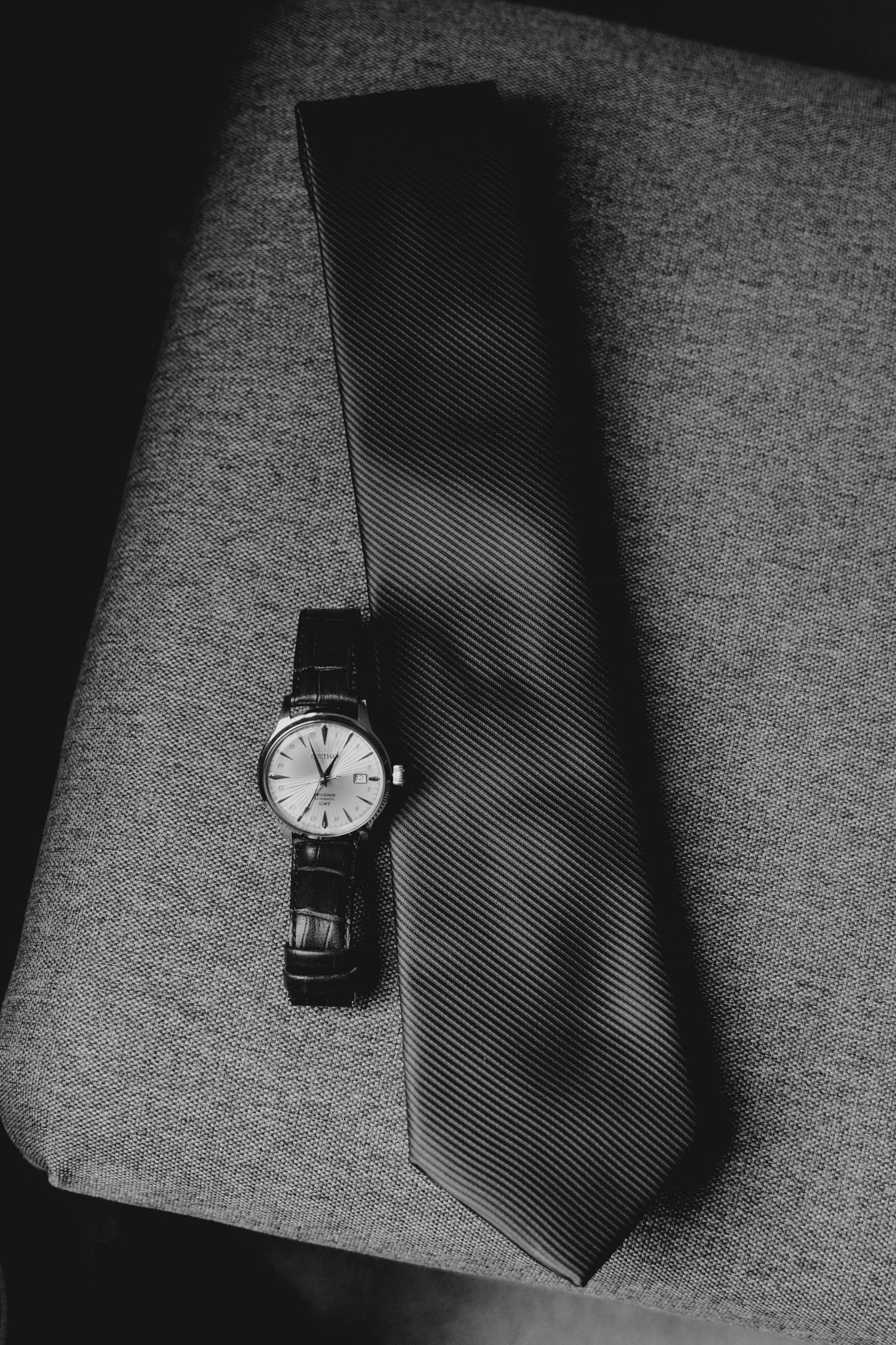 Black and white photo of a classic wristwatch resting on a striped necktie on a gray chair.