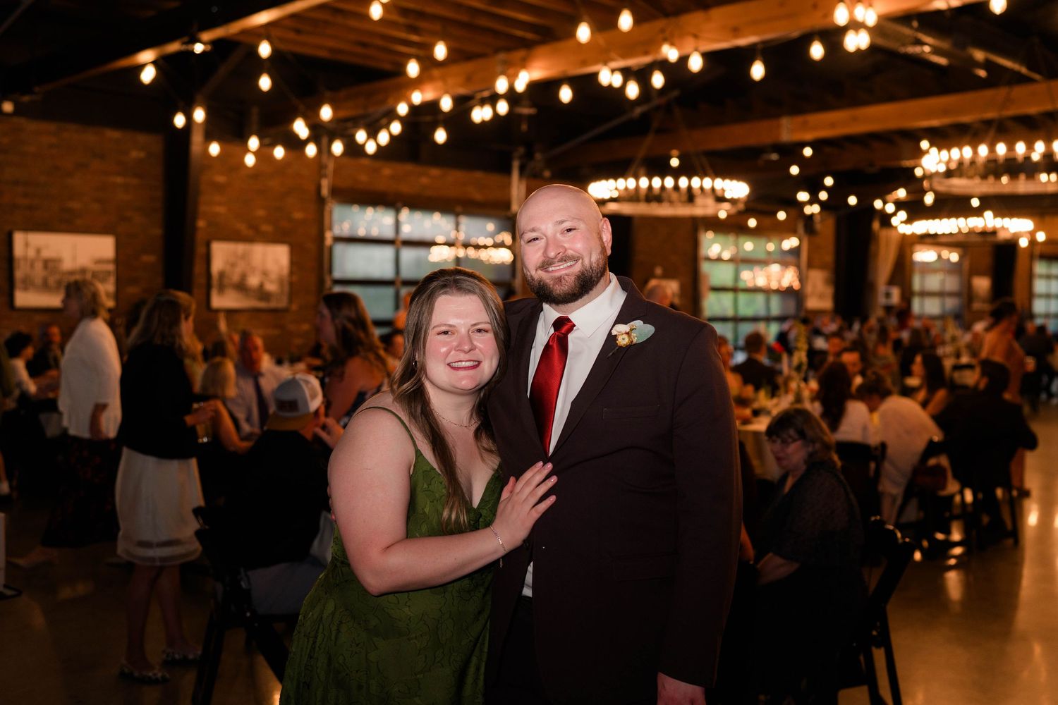 Smiling couple posing together at wedding reception with string lights and brick walls behind them.