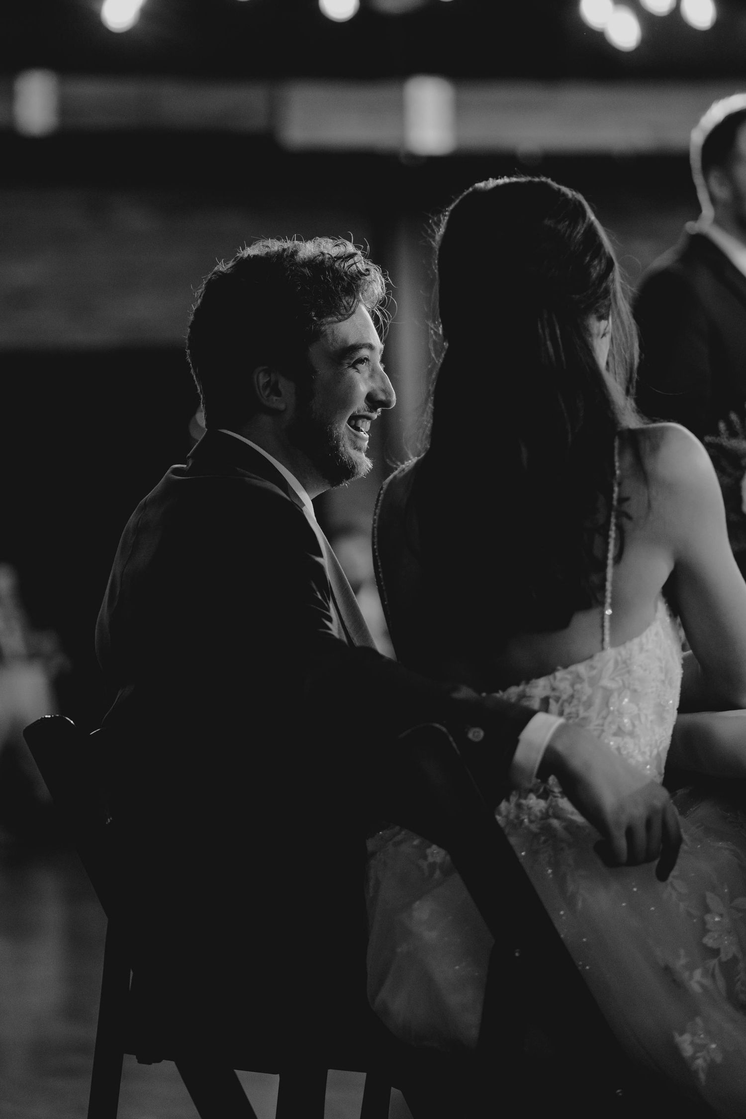 Black and white photo of smiling groom gazing at bride during intimate wedding reception moment.